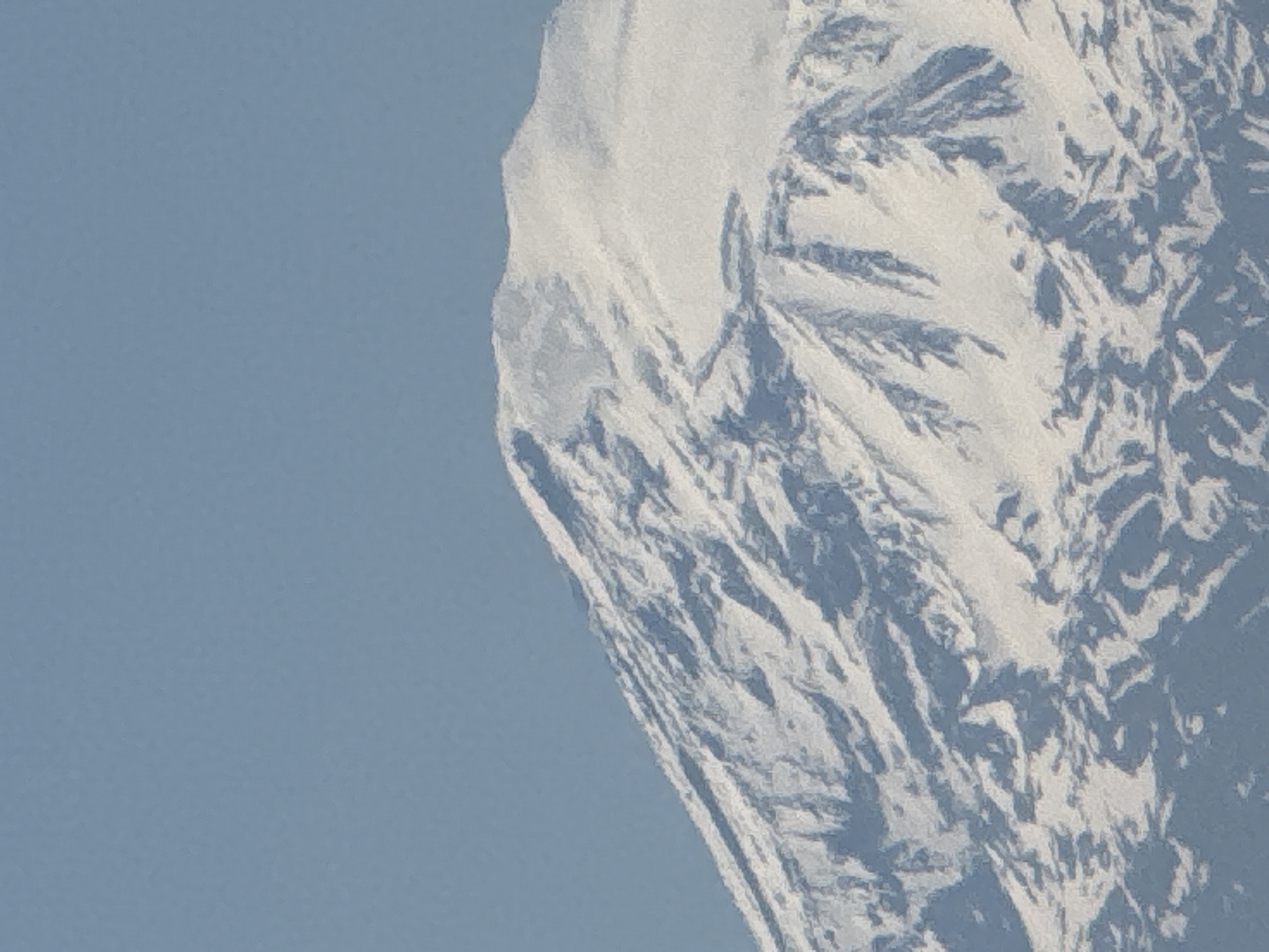 The image displays a close-up view of a majestic, snow-covered mountain or a significant portion of a large rock face. The primary subject is this natural formation, which occupies the right half and extends to the top and bottom edges of the frame. The left side of the image is dominated by a clear, unblemished light blue sky, indicating bright weather conditions with no visible clouds.
The mountain's surface is intricately textured with white snow, exhibiting varied patterns of light and shadow that highlight its rugged topography. Darker blue and grey tones are visible within the snow, suggesting areas of shade, exposed rock, or deeper crevasses. The intricate details of the snow and rock formations create a sense of immense scale and geological complexity. No human activity, structures, or living beings are visible, emphasizing the raw, untamed nature of the environment.
Notable details include the crisp delineation between the mountain and the sky, suggesting clear atmospheric conditions. The lighting indicates it is daytime, with strong directional light creating sharp contrasts across the snow-covered slopes. The overall impression is one of a remote, high-altitude landscape. There is no visible text in the image.