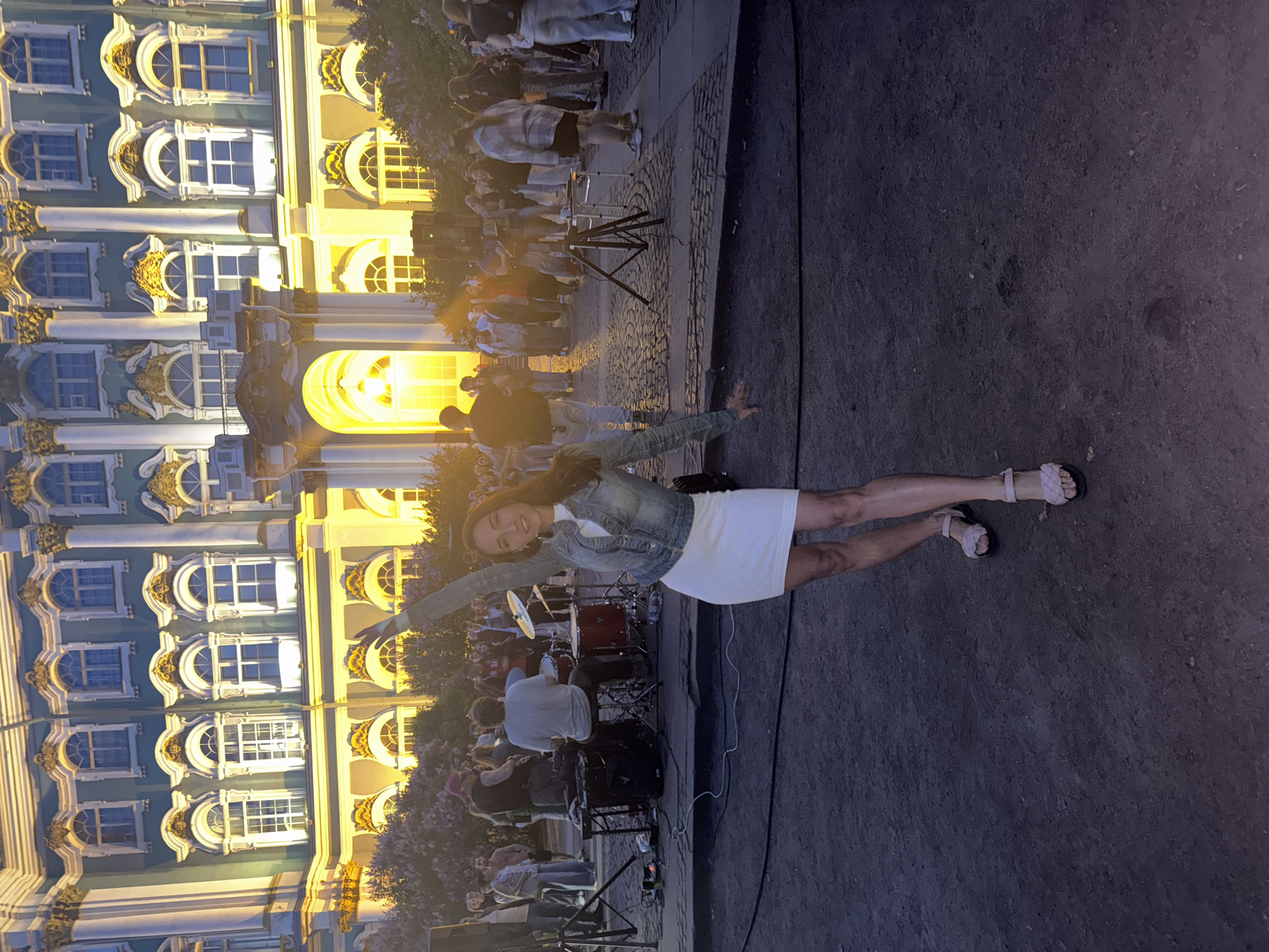 This image depicts an outdoor evening event in Saint Petersburg, Russia, with the prominent backdrop of a grand, ornately decorated building, which strongly resembles the Winter Palace (Hermitage Museum) due to its distinctive pale green/blue and white facade with golden architectural details and numerous arched windows.
In the foreground, a woman with dark hair and a bright smile poses for the camera, her left arm outstretched. She is wearing a white dress, a denim jacket, and light-colored braided sandals. The ground beneath her is a dark paved surface.
Behind the woman, a live music performance is taking place. A drummer is seated at a visible drum kit, and other band members and equipment, such as speakers and stands, are present. A crowd of people is gathered beyond the performers, watching the event.
The scene is set at dusk or early evening, evidenced by the dark ambient light and the dramatic illumination of the building, which glows with warm, golden light emanating from its windows. This artificial lighting highlights the intricate architectural features and creates a festive atmosphere. The overall mood appears lively and celebratory. No discernible text is visible within the image.
