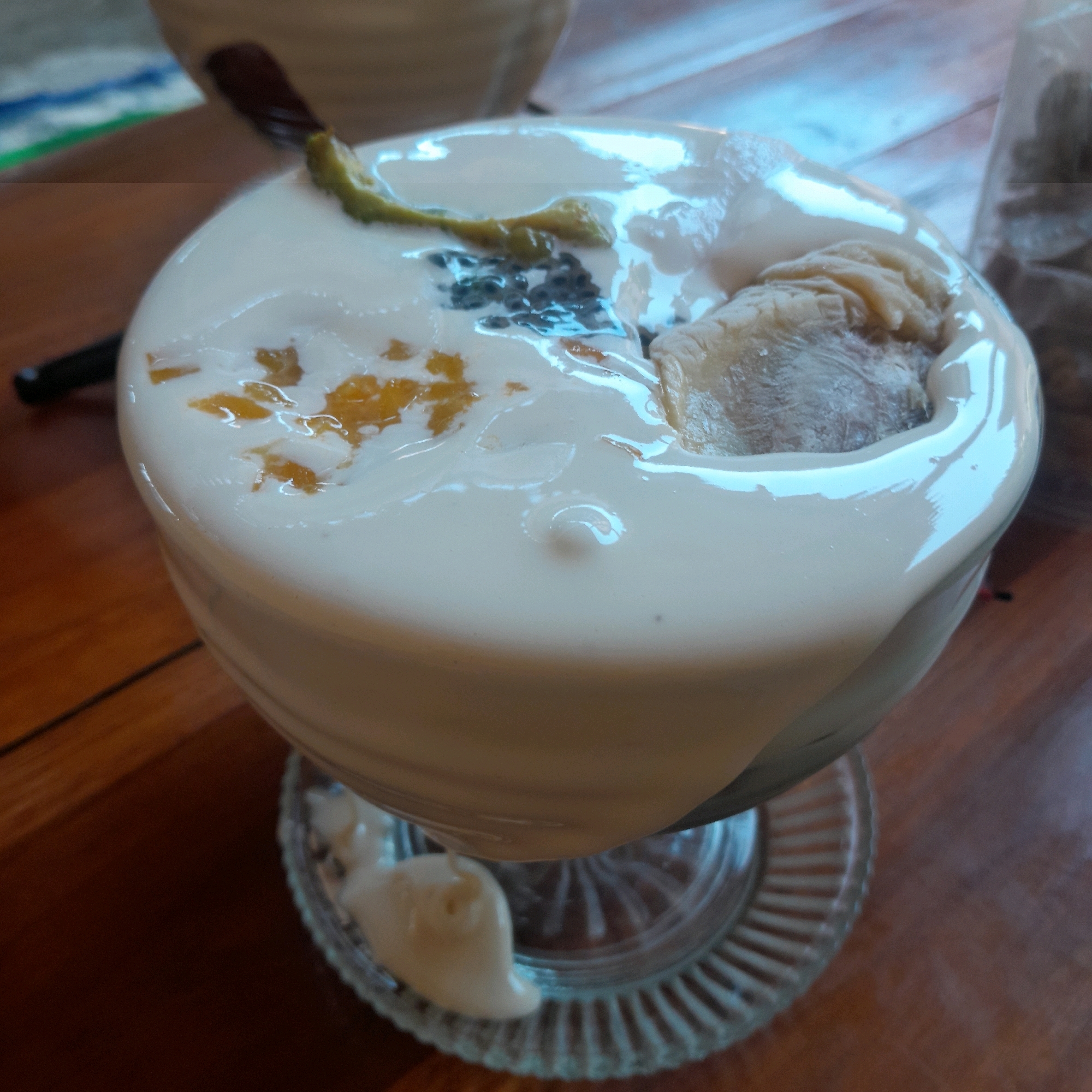 The image shows a close-up view of a dessert in a glass bowl. The dessert appears to be a creamy white substance, possibly yogurt or pudding, with chunks of fruit, likely mango, mixed in. There are also some black seeds and a sliver of green peel on top. A piece of what looks like ice cream or solidified cream is partially submerged in the dessert. The bowl is resting on a decorative glass plate. The background is a wooden surface, suggesting the dessert is on a table. A dark object, possibly a spoon or a utensil, is partially visible to the left of the bowl. The overall impression is that of a refreshing and likely sweet treat. The scene is likely a cafe or restaurant in Terung Wetan, Indonesia, given the context provided.