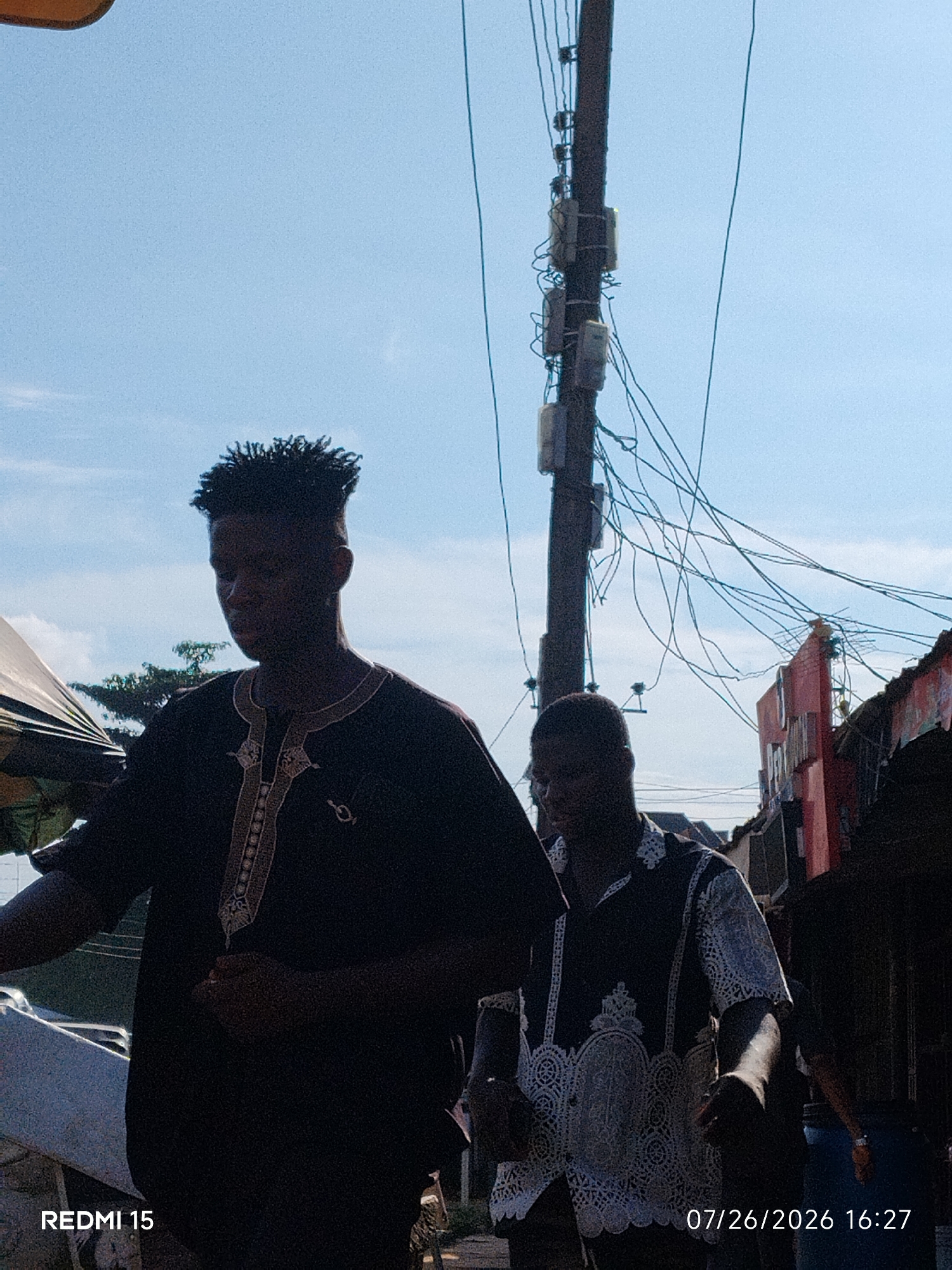 This is a medium shot, outdoor, daylight photograph of two young men walking. The man in the foreground, on the left, is wearing a dark, traditional African garment with gold embroidery around the neckline and down the front. He has short, dark, textured hair. The man behind him, on the right, is wearing a patterned shirt with white decorative elements. They are walking on a street with a tall wooden utility pole covered in electrical wires and boxes in the background, along with buildings and trees. The sky is blue with some clouds. The photograph was taken in Umuode, Nigeria, as indicated by the timestamp and device information in the lower corners.