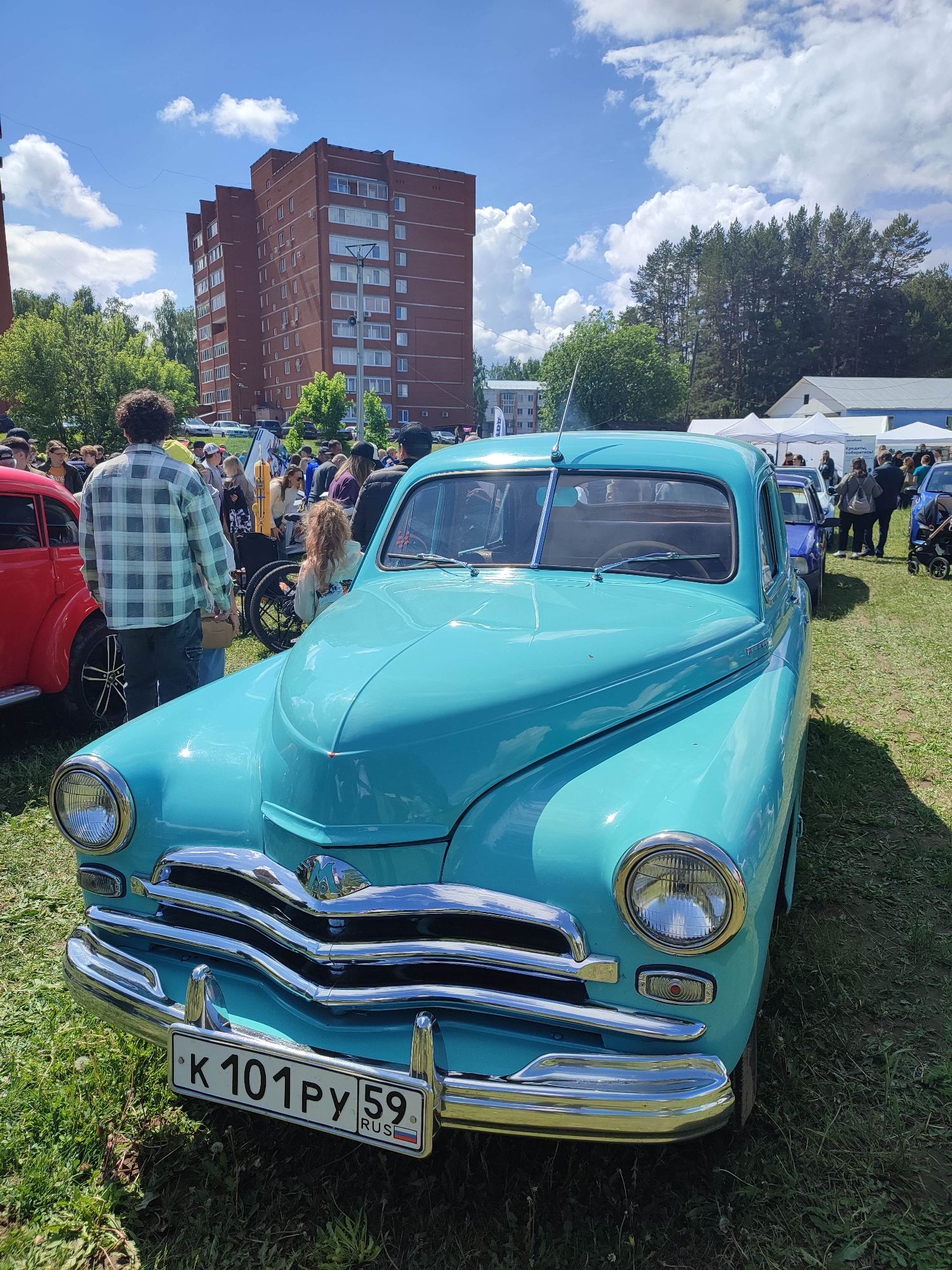 The image shows a bright turquoise vintage car on a grassy field. The car has a Russian license plate reading "K 101 PY 59 RUS". The chrome grille and bumper are prominently displayed. Behind the car, there is a gathering of people, suggesting an outdoor event. A red car is partially visible to the left. In the background, there is a multi-story brick building, trees, and some white tents. The location appears to be a public outdoor space, possibly a park or event area. The sky is blue with scattered white clouds.