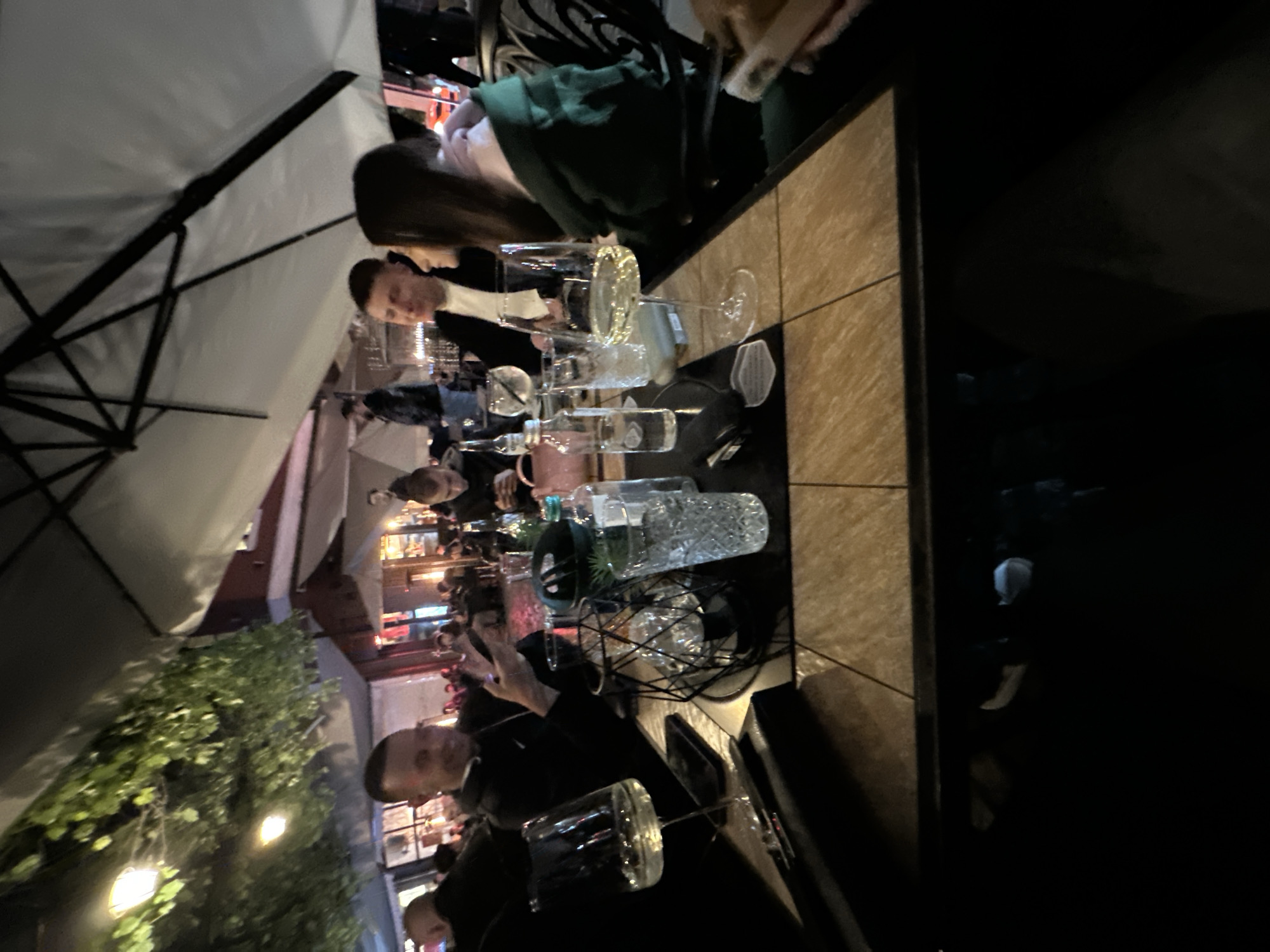 A group of people are sitting at an outdoor table under a large, white umbrella at night. The table is covered with glasses, dishes, and other items. There are trees and lights in the background. The location is likely in the city of Plovdiv, Bulgaria.