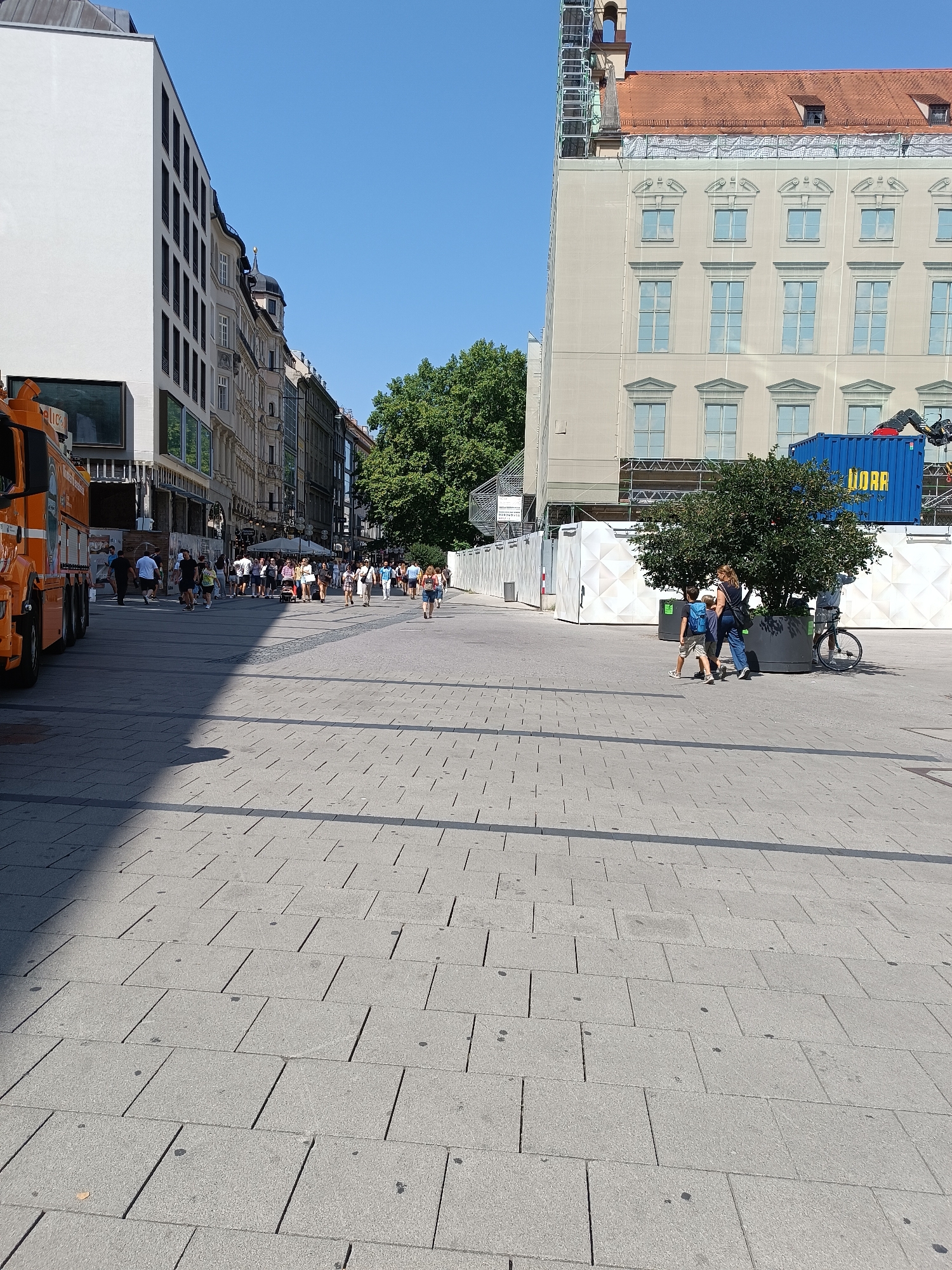 The image captures a sunny day on a pedestrianized street in Munich, Germany. The street is paved with grey interlocking tiles. On the left, a white modern building with dark-framed windows stands tall, partially obscuring older buildings behind it. An orange truck, possibly a food truck, is parked on the far left.
In the background, the street is lined with traditional European buildings, some with ornate facades and tiled roofs. Construction scaffolding is visible on a large, pale building on the right, which is covered in a protective white tarp. A blue shipping container labeled "LIDAR" is also present near the construction site.
A crowd of people are walking along the street, some in small groups, others individually. They are dressed in casual summer clothing. In the mid-ground on the right, a woman with a backpack and a young boy, also with a backpack, are walking together. A bicycle is parked near them. Lush green trees are scattered throughout the scene, providing shade. The sky is a clear, bright blue. The overall impression is one of a vibrant urban environment on a warm day.