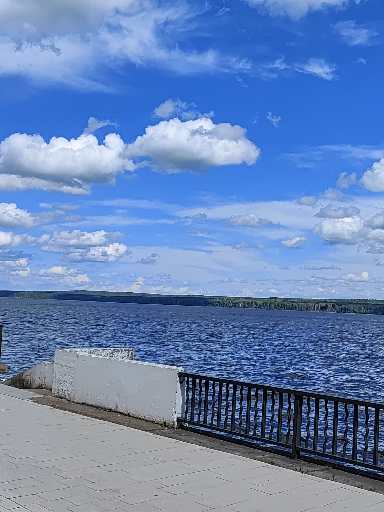The image depicts a scenic view of the Kama River in Chaikovsky, Russia. In the foreground, a paved walkway runs alongside the river, bordered by a black metal railing. Adjacent to the walkway is a low, white concrete wall. The river itself is a deep blue, with visible ripples on its surface. In the distance, a lush green forest lines the opposite bank of the river, forming a dark, horizontal band against the horizon. The sky above is a vibrant blue, dotted with numerous white, fluffy cumulus clouds. The overall impression is one of a tranquil and picturesque waterfront setting on a clear, sunny day.