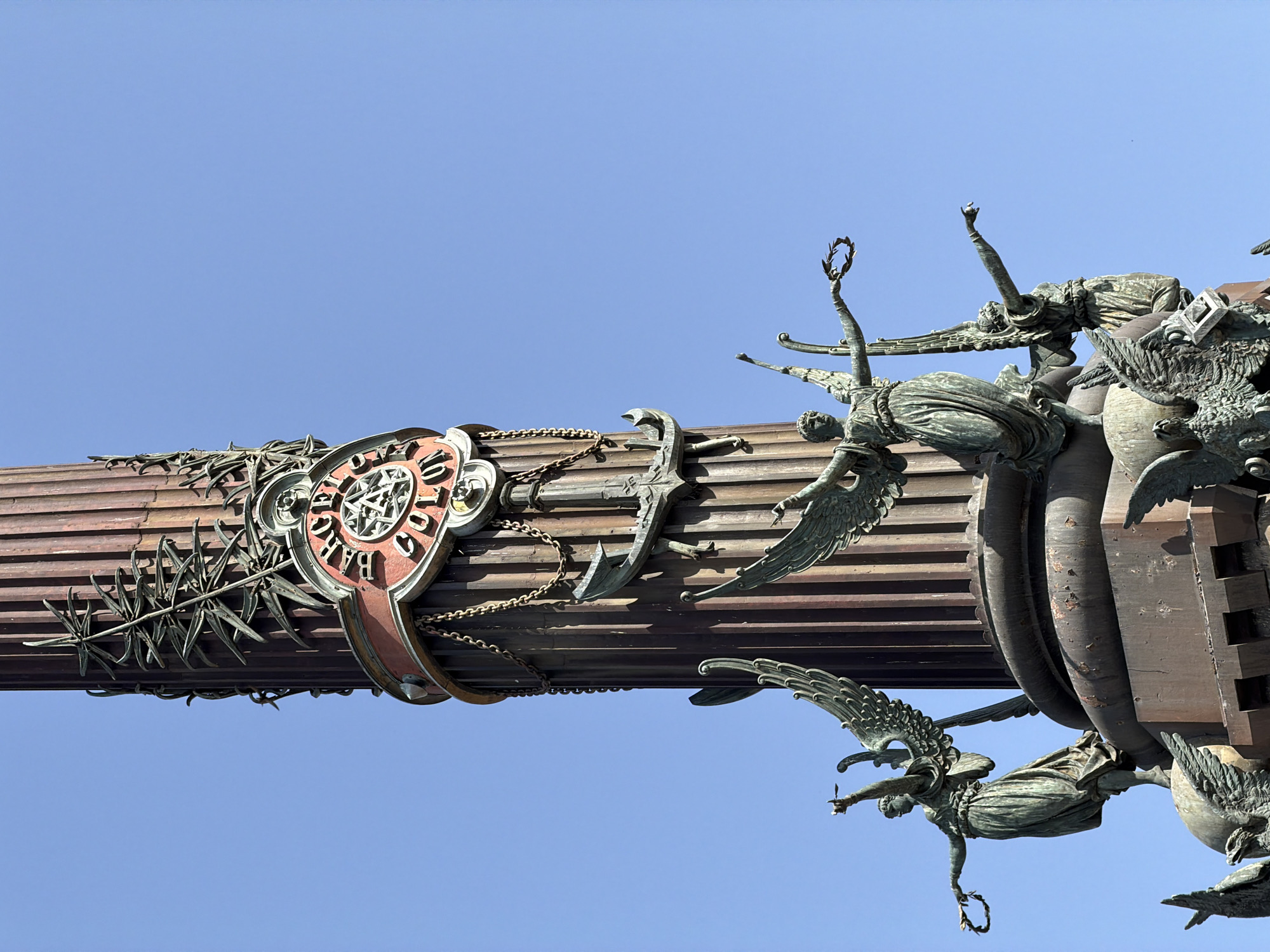 The image shows the Monument to Christopher Columbus, located at the bottom of Las Ramblas in Barcelona, Spain. It is a tall, fluted column made of bronze and stone, topped by a statue of Christopher Columbus. The monument is decorated with various sculptures and reliefs, including a statue of Columbus pointing towards the sea, and figures of winged Victories holding laurel wreaths. In the foreground, several bronze sculptures of winged figures are visible, some holding wreaths and others pointing. The monument stands against a clear blue sky.