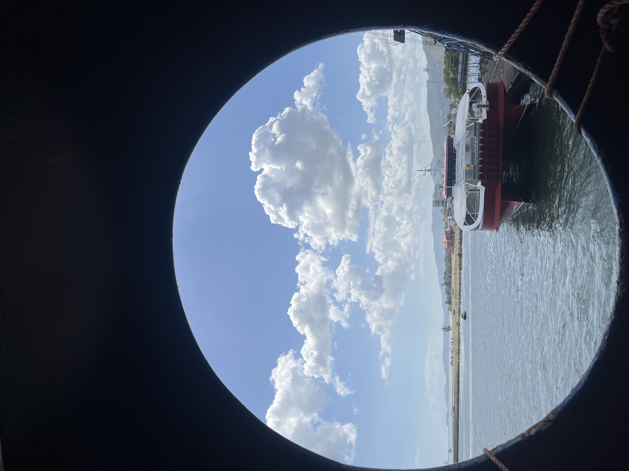 The image captures a vibrant daytime scene viewed through a dark, circular porthole, presumably from within a vessel. The overall perspective is of a harbor or a dockside area.
The top portion of the view through the porthole reveals a clear, bright blue sky dotted with numerous fluffy, white cumulus clouds. These clouds vary in size and shape, with some appearing large and prominent.
Below the sky, a body of calm water, likely a sea or a large river, occupies the middle and lower sections of the circular frame. Gentle ripples are visible on its surface. To the right, within the porthole's view, the bow section of a large, docked vessel is prominent. The ship features a dark red hull with a white superstructure and deck, and appears to be a working ship, possibly a fishing trawler or a smaller cargo vessel.
Across the water, a distant shoreline stretches horizontally, featuring an area that appears industrial or port-related, with various structures and possibly cranes or buildings. Further in the background, a range of mountains or hills rises, partially obscured by a slight atmospheric haze.
The dark, thick frame of the porthole forms the immediate foreground, encompassing the visible scene. A braided rope is visible along the inner edge of the porthole frame in the lower right, suggesting it's tied to the vessel from which the photo was taken. The scene is brightly lit, indicating midday or early afternoon conditions with good visibility. There is no visible text in the image.