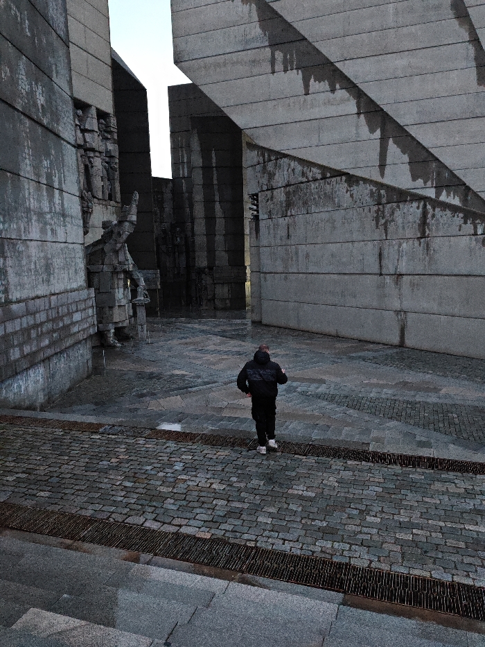 The image captures an outdoor scene within the Monument to 1300 Years of Bulgaria in Shumen, Bulgaria. A single male individual is depicted from behind, walking rightward across a wet, grey cobblestone path. He is dressed in a dark hooded jacket, dark trousers, and white athletic footwear.
The environment is dominated by massive, brutalist-style concrete structures forming angular walls and passageways. These structures exhibit a rough, block-like texture and display dark vertical streaks, indicating water runoff and weathering. To the left, a large, geometric concrete sculpture of a seated or kneeling human figure is partially visible. The ground in the foreground consists of grey concrete steps, transitioning to metal grates, then the cobblestone path, and further into the monument, a darker, wet paved area. The sky above the structures appears light and overcast. Patches of standing water are visible on the ground surfaces.