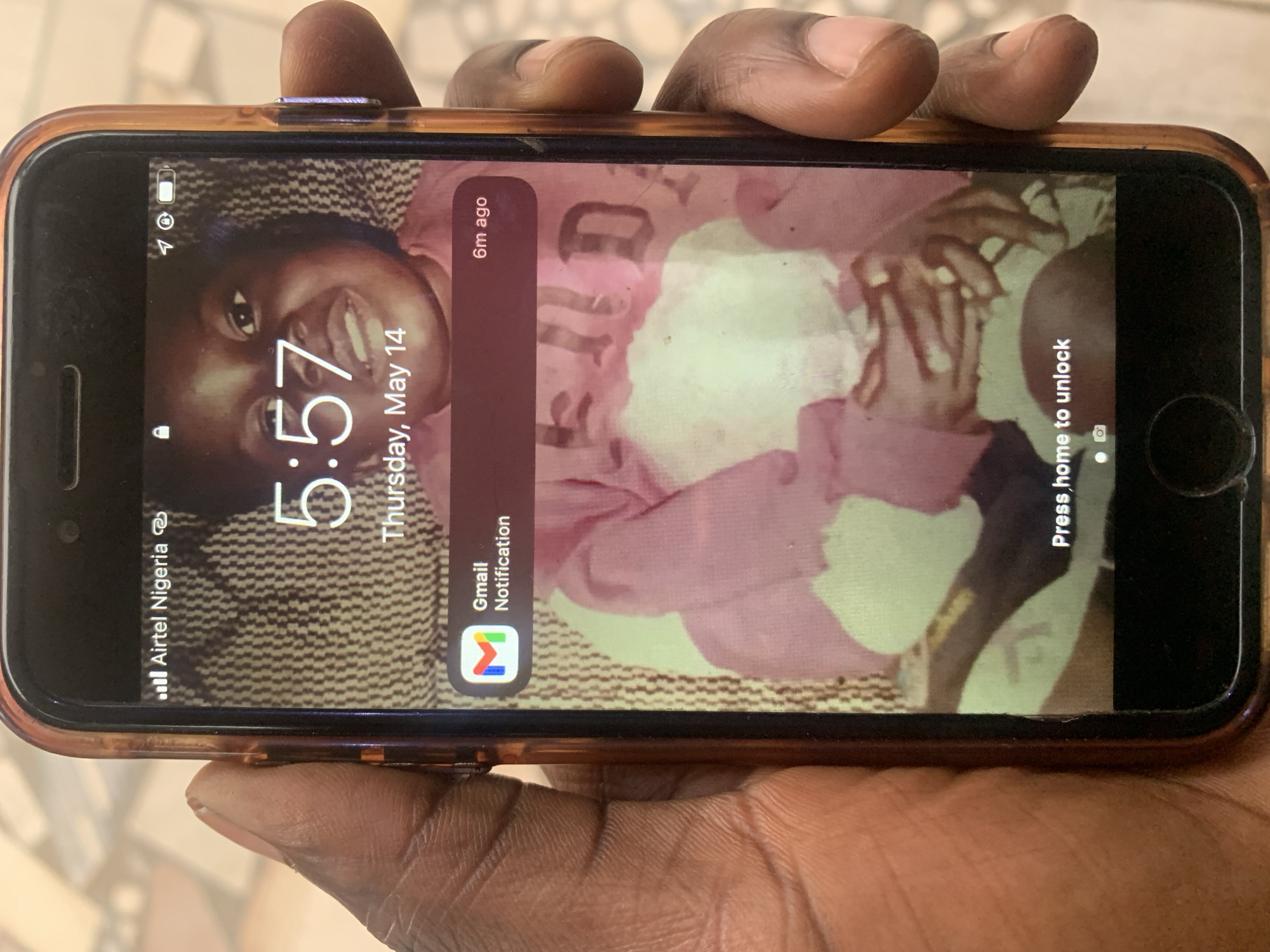 A hand holds an iPhone displaying a lock screen. The time is 5:57 on Thursday, May 14th. There is a Gmail notification. The phone shows the carrier Airtel Nigeria. The wallpaper is a picture of a smiling black woman. The location is Kabong, Nigeria.