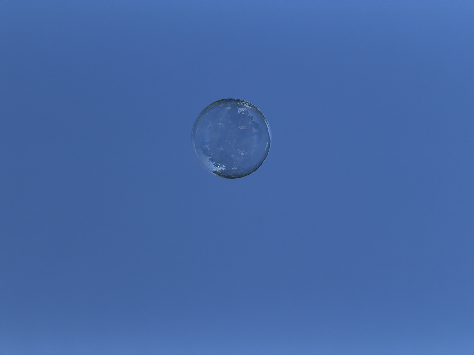 A solitary bubble drifts against a clear blue sky. The bubble is transparent and spherical, reflecting the surrounding environment, which appears to be trees or buildings. The background is a uniform expanse of blue, indicating an outdoor setting. The image was captured in Chaikovsky, Russia.