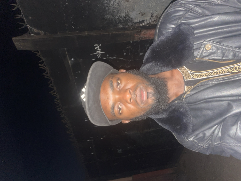 A dark-skinned man with a full beard poses in a gray baseball cap. He wears a black leather jacket with a black fur collar over a shirt with gold embroidery around the neckline. The setting appears to be nighttime outdoors, with a dark structure topped with barbed wire visible in the background. Writing is visible on the structure.