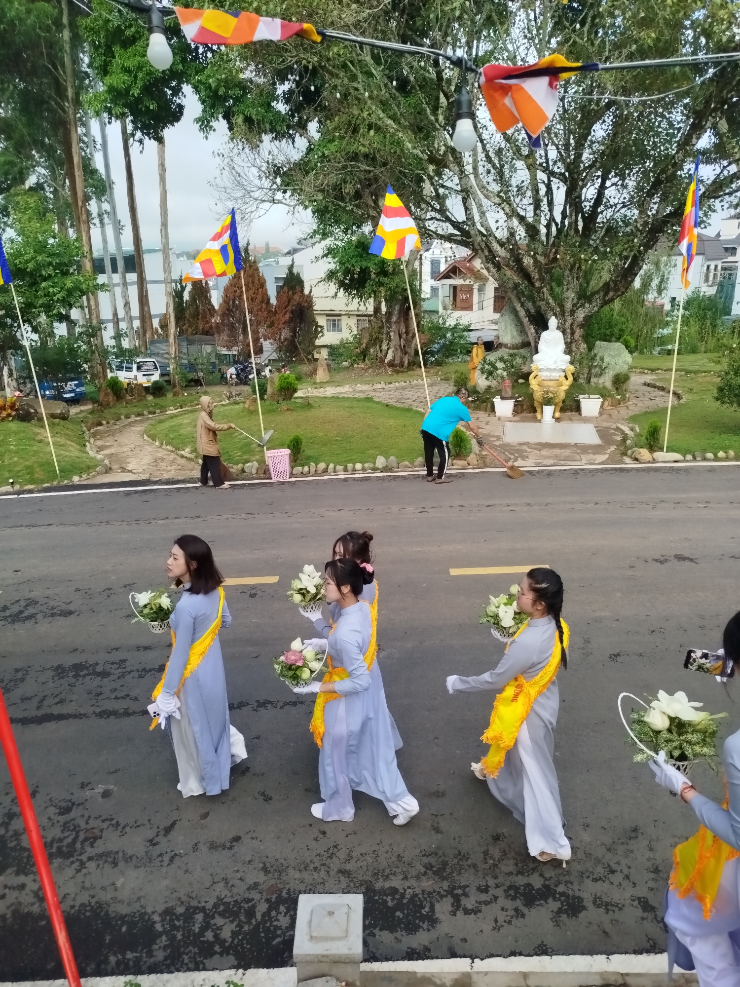 The image shows a street scene with people involved in various activities, likely in Da Lat, Vietnam. In the foreground, several young women are walking along the road carrying baskets of flowers. They are dressed in traditional blue clothing with yellow sashes. In the background, two people are cleaning the area around a statue of Buddha that is on a raised platform surrounded by greenery. The street appears to be asphalt and has yellow lines painted on it. Trees line the street, and Buddhist flags are hung overhead. The overall impression is a religious procession or festival preparation.