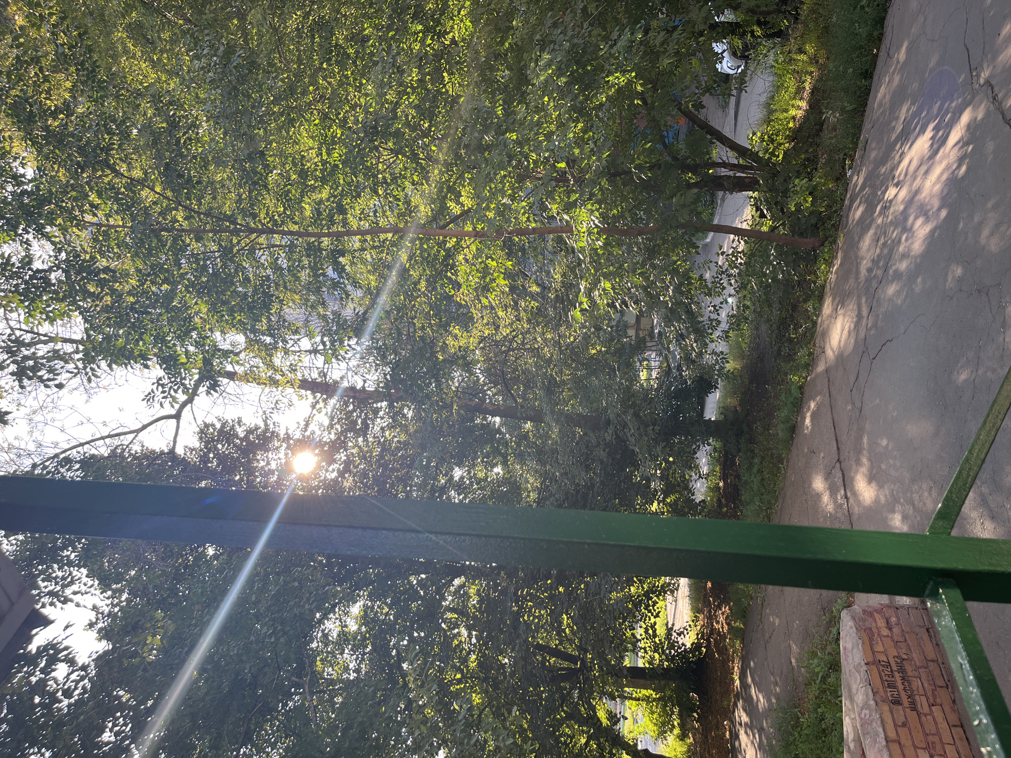 The image captures an outdoor scene, likely a residential area, under bright sunlight filtering through trees. A prominent green metal post is in the foreground, partially obscuring a pathway made of cracked concrete. The pathway is bordered by a low brick structure with some writing or graffiti on it. Lush green trees with dense foliage dominate the background, with sunlight creating strong lens flares and dappled shadows on the ground. In the distance, buildings and a white car are visible, suggesting an urban or suburban environment. The overall impression is one of a sunny day in a green, possibly well-established, neighborhood.