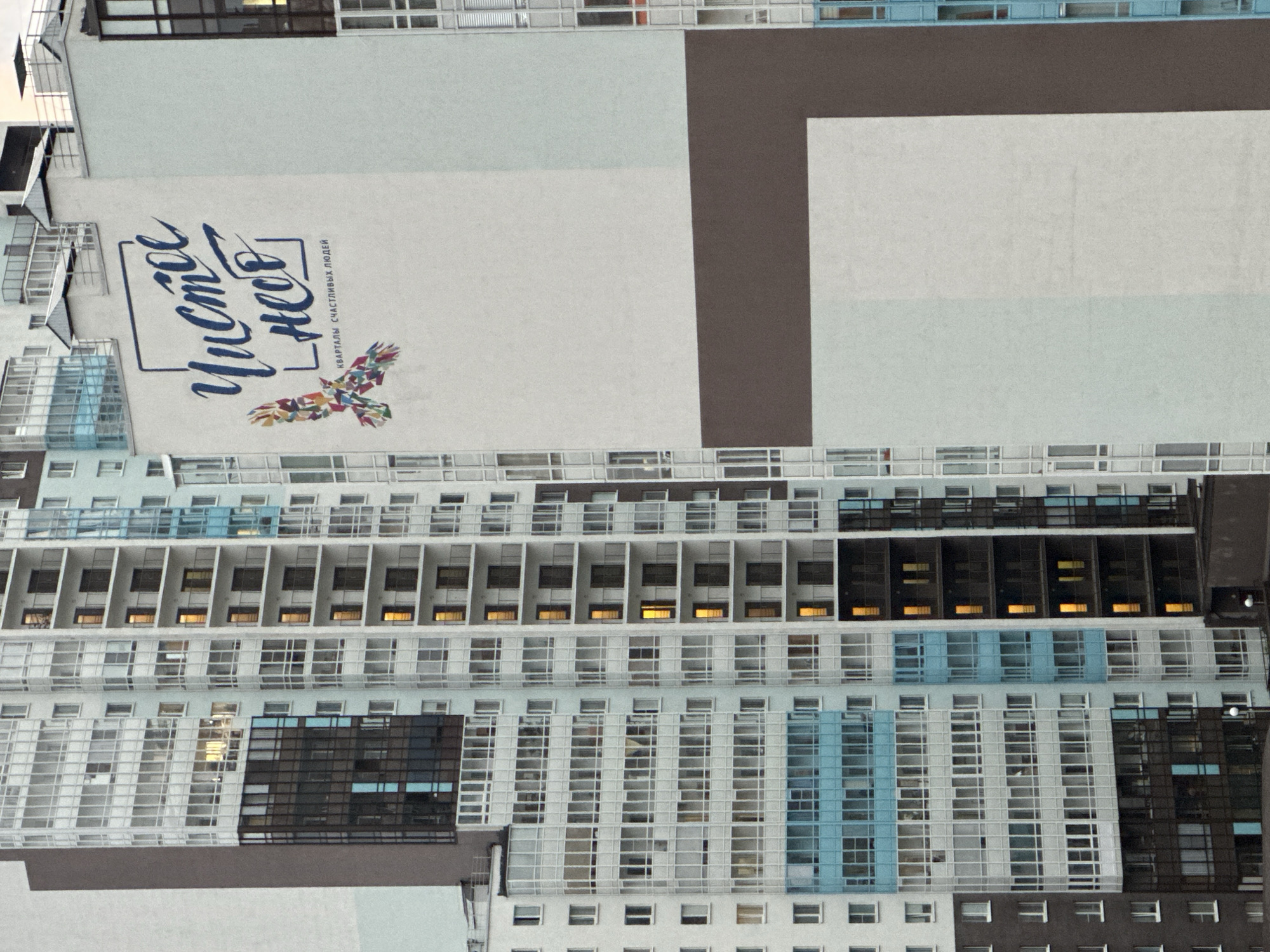 The image shows a close-up of a residential building in Saint Petersburg, Russia. The building is tall and modern, with many windows and balconies. One side of the building features a large mural with the text "Чистое небо" (Pure Sky) and an image of a bird made of colorful geometric shapes. Below the text, it reads "КВАРТАЛЫ СЧАСТЛИВЫХ ЛЮДЕЙ" (Quarters of Happy People). The mural is painted on a white section of the building, contrasted with a large dark brown horizontal band below it. The rest of the building is composed of white and light blue sections, with numerous windows reflecting the sky and interior lights. The sky is overcast. No people are visible in the image. The overall scene depicts a residential complex in an urban environment.