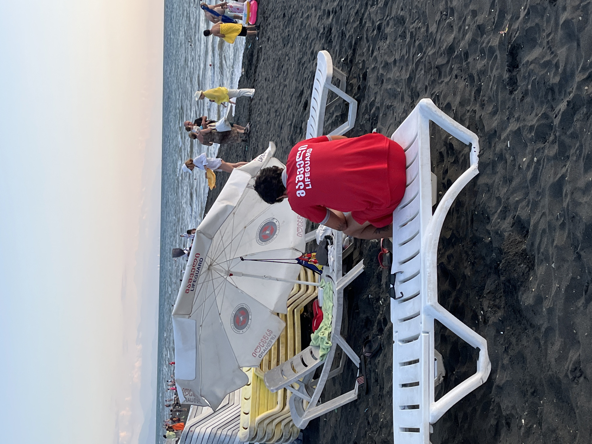 The image depicts a black sand beach in Ureki, Georgia, on a clear day with a calm sea. In the foreground, a lifeguard wearing a red t-shirt with white lettering that reads "LIFEGUARD" in both Georgian and English sits on a white plastic lounge chair. The lifeguard is facing away from the camera, looking towards the ocean.
Scattered around the beach are several white plastic lounge chairs, some stacked and others in use. A white beach umbrella with a logo is partially visible, casting a shadow over some of the chairs. In the background, a few people are visible near the water, some wading, while others are walking along the shore or sitting on the sand. The overall scene suggests a relaxed atmosphere at a resort beach.