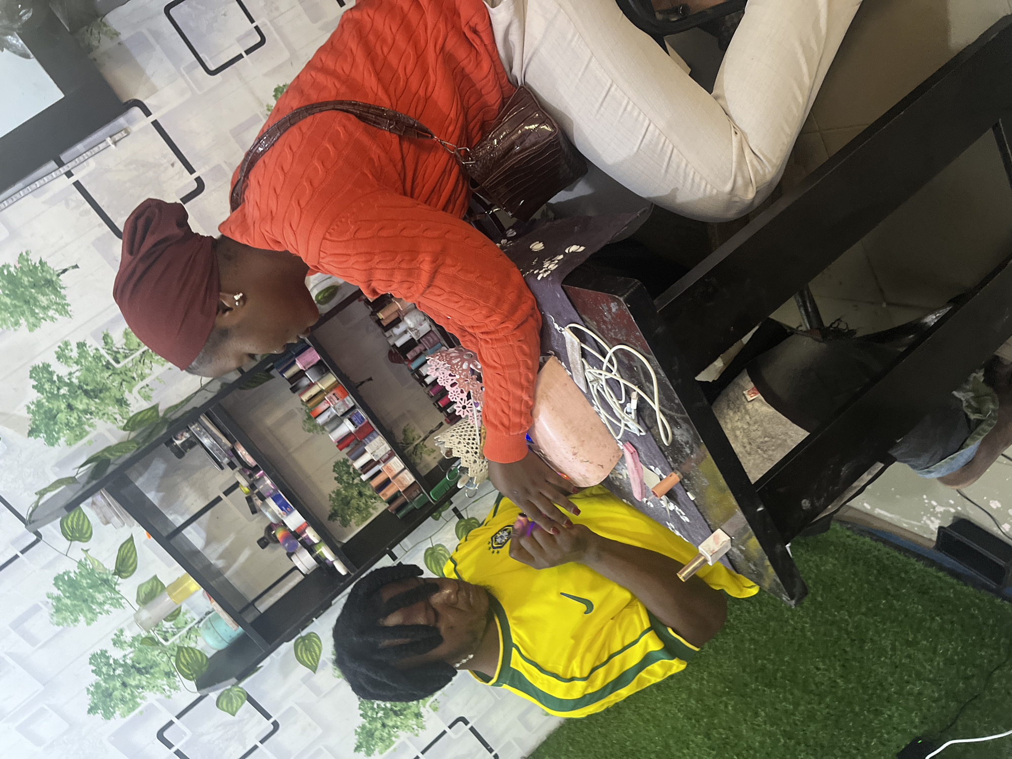 In a well-lit nail salon in Jos, Nigeria, a nail technician with dreadlocks, wearing a yellow and green jersey, applies nail polish to a client's fingernails. The client, a woman with a maroon headscarf and an orange cable-knit sweater, is seated at a black table adorned with various nail polish bottles and tools, including a pink nail buffer. A brown textured purse hangs from the client's shoulder. The salon's walls are decorated with a wallpaper featuring a repeating pattern of green trees against a white background with black square outlines. A black shelf unit is visible behind the individuals, stocked with numerous nail polish bottles in an array of colors. The floor is covered with artificial turf in the foreground, transitioning to tiled flooring in the background, indicating a professional setup within the city of Jos.