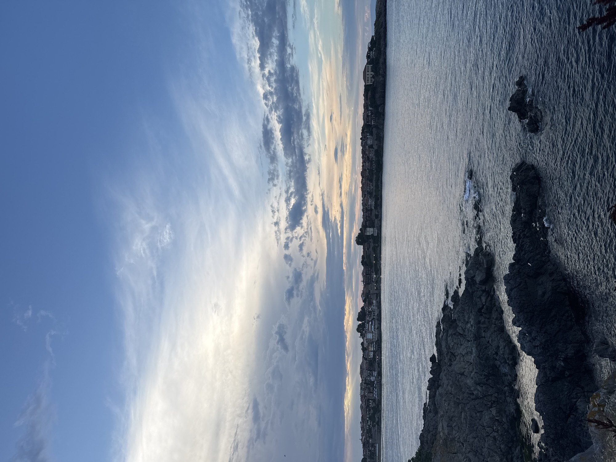 This is a landscape photograph of the sea and coastline of Sozopol, Bulgaria. In the foreground, there are dark, jagged rocks and the calm, dark blue sea. The middle ground features the coastline with a town of red-roofed buildings and trees. The sky above is a mix of blue and white clouds, with some lighter clouds near the horizon suggesting the setting or rising sun. The overall lighting is soft and diffused, indicative of twilight. There are no people or visible actions in the scene.