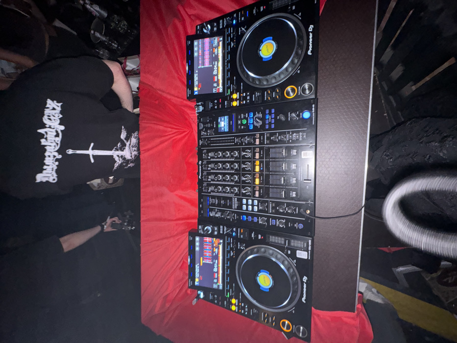 The image displays a dimly lit indoor event, likely a nightclub or concert venue, viewed from a slightly elevated perspective towards a performance area. The foreground is dominated by a professional DJ setup consisting of two black Pioneer DJ CDJ-3000 multi-players flanking a central Pioneer DJ DJM-A9 mixer, all resting on a dark, textured table. Both CDJ-3000 units are active, with illuminated jog wheel centers showing blue and yellow concentric circles, and their respective screens displaying audio waveforms and track information; the left screen indicates a time of 02:51, and the right screen shows 03:24. Various buttons and faders on all three devices are illuminated with blue, white, and amber lights. A black audio cable is visible, extending from the mixer towards the bottom of the frame. Directly behind the equipment, a red fabric or covering is draped across a surface. In the immediate bottom-center, a blurred, metallic coiled object, possibly a cable, is partially visible.
In the background, several individuals are present within a dark environment. One person, observed from behind, is wearing a black t-shirt with a white graphic depicting a sword plunged into a landscape, surmounted by stylized text (appearing as "Burzum" or similar in a black metal font). To the left of this individual, an arm is visible holding a small, dark object. Other indistinct figures and blurred shapes suggest a crowd in attendance. The specific location within the city of Moscow, Russia, cannot be determined from the visual information presented in the image.