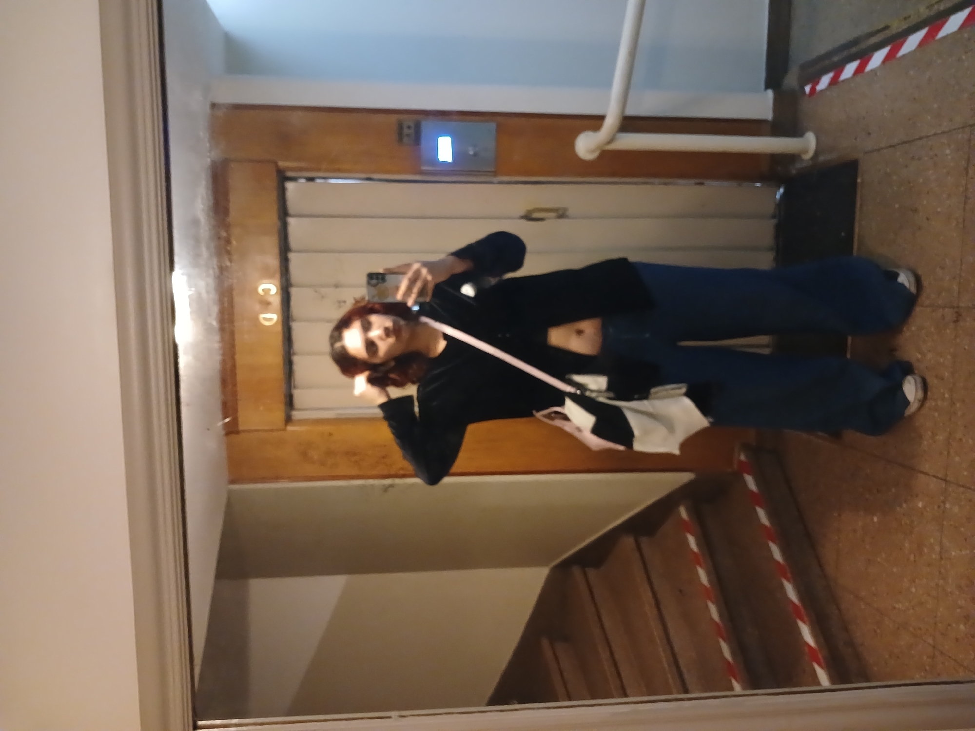 A mirror reflects a person with dark hair and a black top taking a selfie. The background shows an interior space with a wooden-paneled elevator door with an illuminated call button, a white handrail, a staircase marked with red and white tape, and a brown tiled floor. The location might be an apartment building in Buenos Aires, Argentina, given the architectural style and general appearance of the space. The person's action is taking a self-portrait photograph in the mirror.
