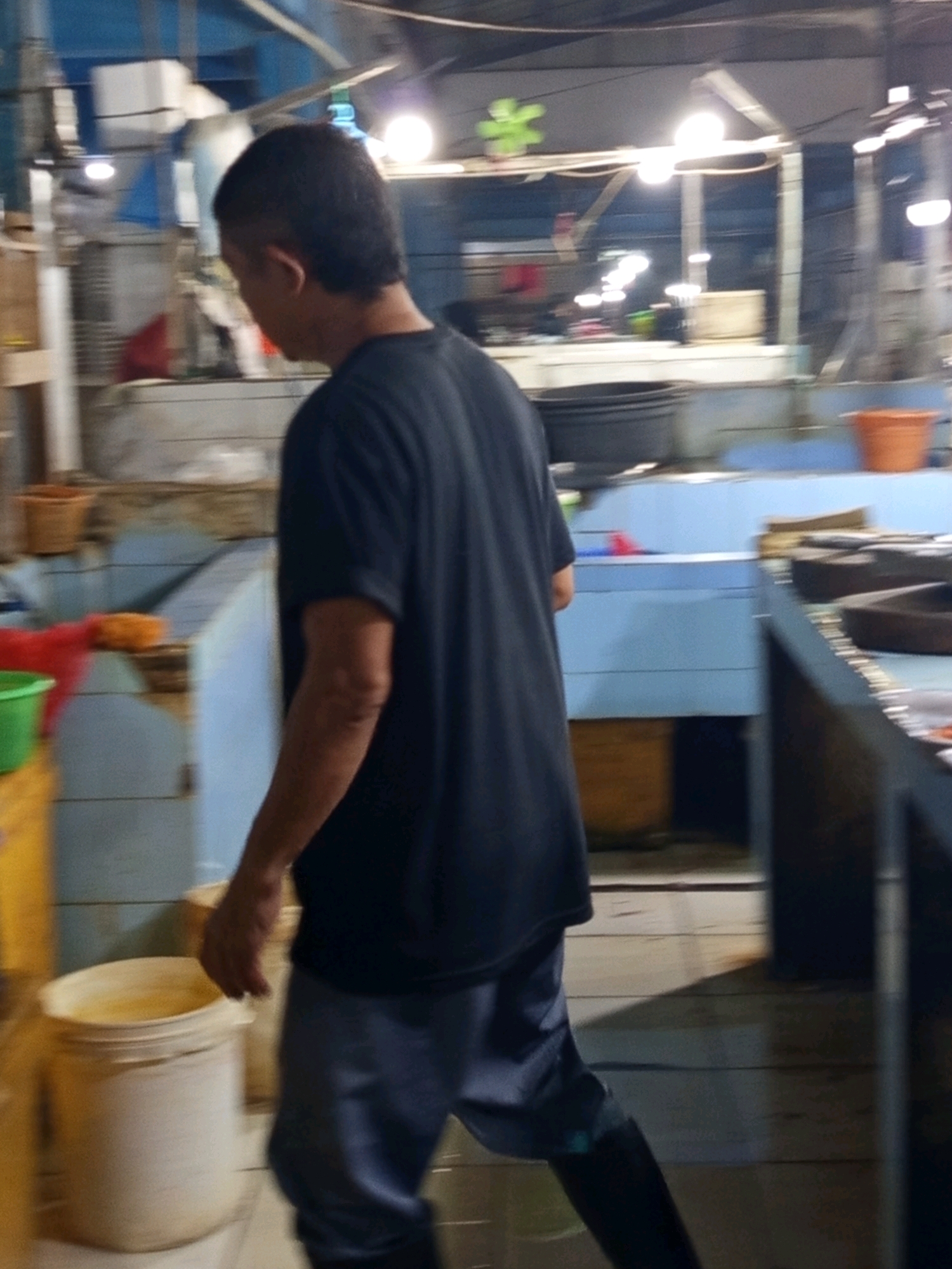The image shows a person wearing a dark t-shirt and blue pants, walking in what appears to be a market or processing facility. The environment is dimly lit with overhead lights visible. There are various containers and equipment in the background, including what looks like large black tubs, a bright orange bucket, and several large white buckets. The floor is made of wood planks. The scene suggests a commercial or industrial setting where goods are being handled or stored. The person's posture indicates they are moving from left to right, away from the viewer.