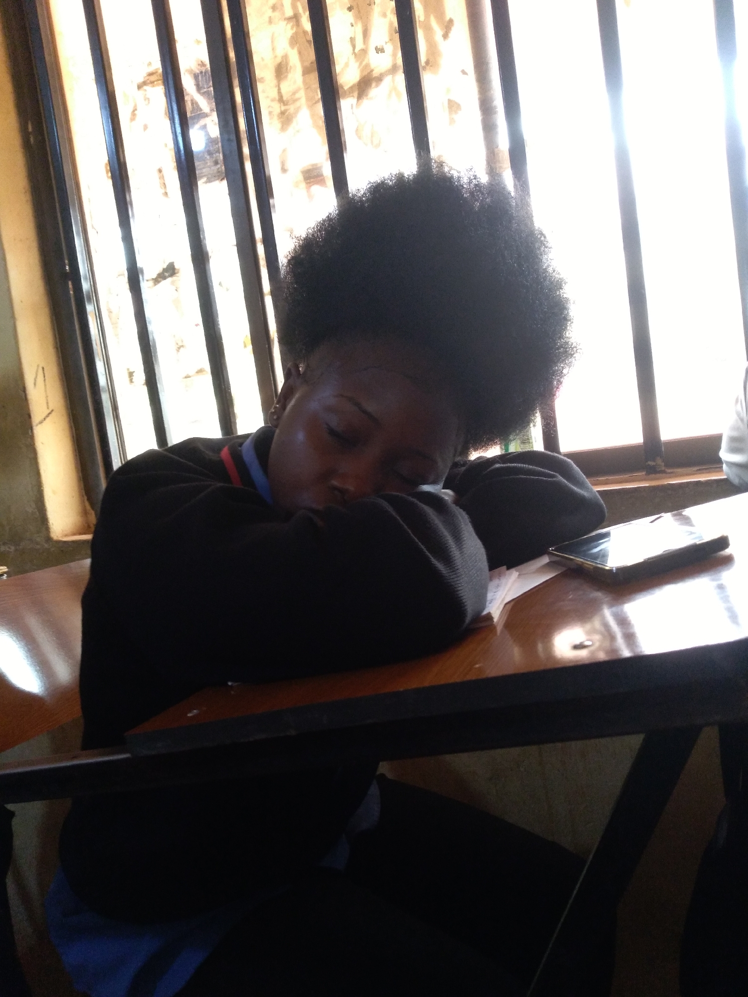 A young Black female, with a large afro hairstyle, is asleep at a wooden desk, her arms crossed and head resting on them. She is wearing a black sweater over a blue collared shirt with a red and blue stripe trim. A smartphone rests on the desk to her right. The background consists of a window with vertical bars, through which bright sunlight streams. The scene is likely a classroom or study environment in Jos, Nigeria, given the light and architectural elements that are common in the region.