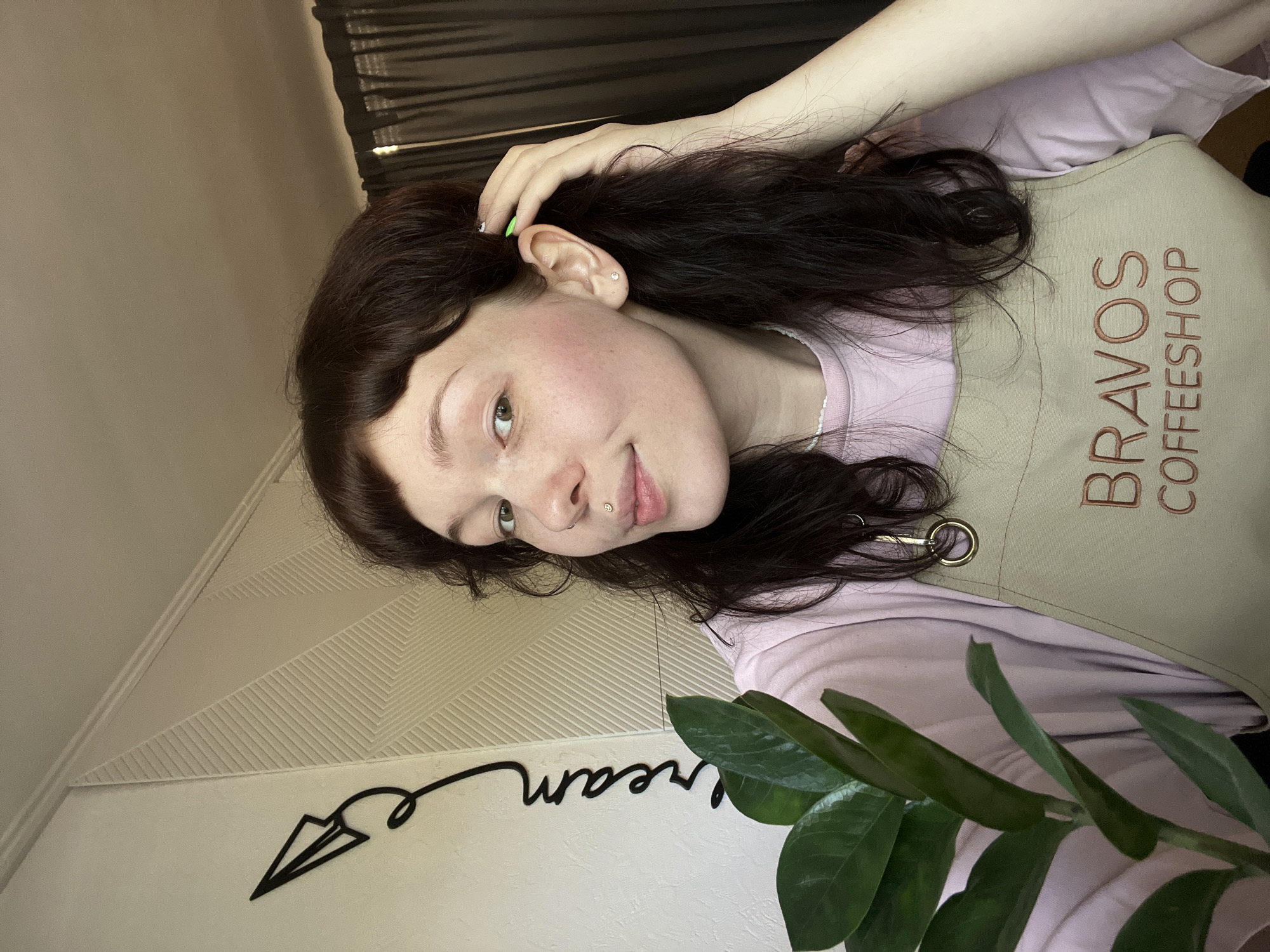 A young woman, likely in her late teens or early twenties, is shown from the chest up. She has dark brown hair with short bangs, light skin with some freckles, and green eyes. She wears a light purple t-shirt and a beige apron with the words "BRAVOS COFFEESHOP" embroidered on it. She has a piercing on her lip and a green fingernail on her left hand, which is resting on her head.
The background features a wall with a geometric pattern and a black decorative item resembling a paper airplane with the word "dream" written in cursive. A plant with green leaves partially obscures the bottom right corner of the image. Behind the woman, dark curtains can be seen.
There is no definitive evidence in the image to confirm the location as Chaikovsky, Russia. The apron suggests she may be an employee of a coffee shop, but there is nothing specific to link it to that location.