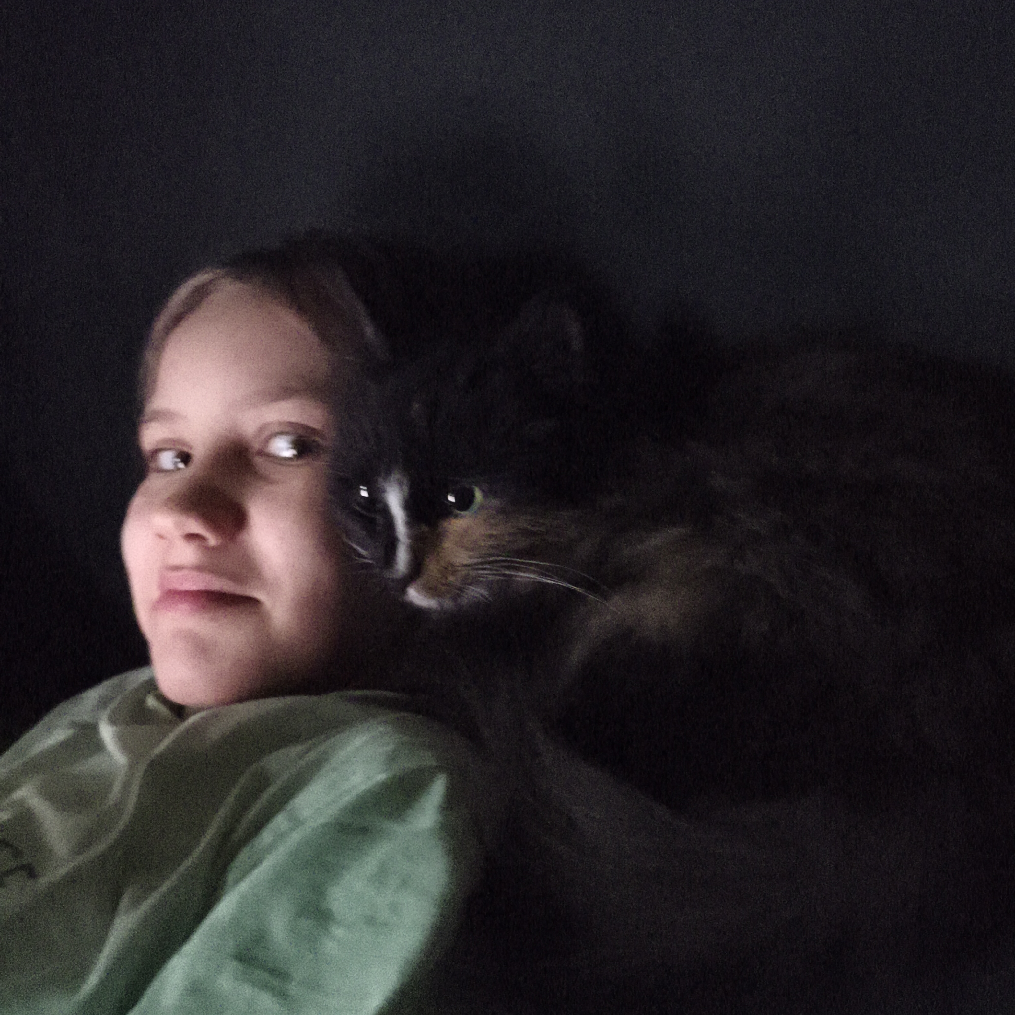 A girl with fair skin, wearing a green top, is lying down with a black and white cat perched on her shoulder. The background is dark, obscuring details about the environment. Location is Unknown, Unknown.