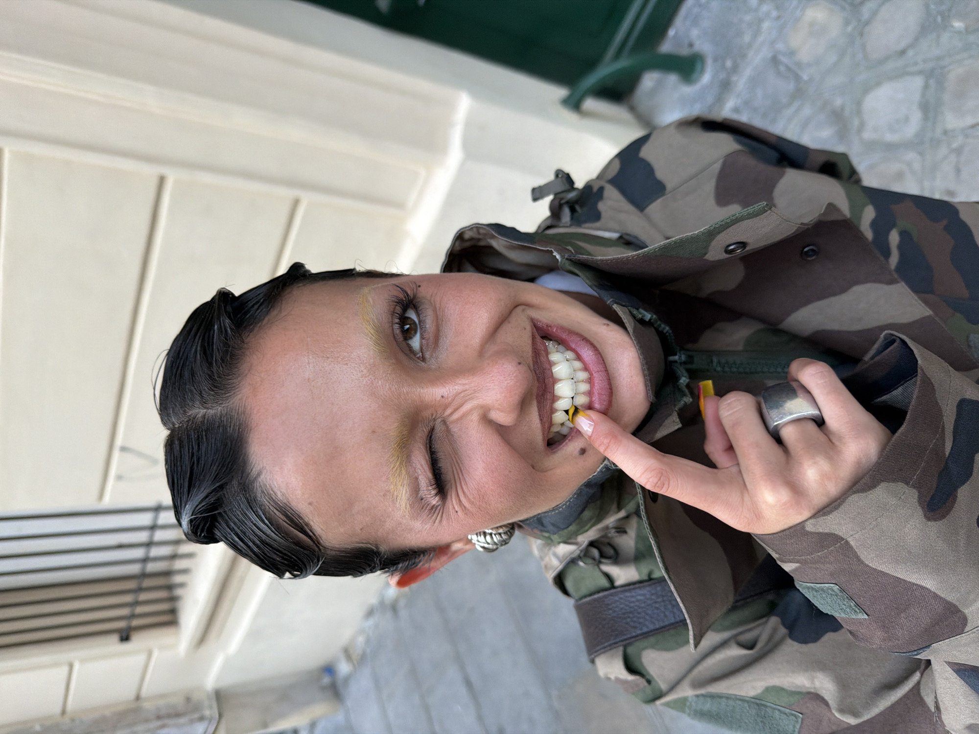 A woman with dark, slicked-back hair, and yellow-dyed eyebrows, wearing a camouflage jacket, is posing, sticking her tongue out with a yellow gummy candy on her tongue. She is winking, and wearing a large silver ring. The background appears to be a city street with building facades and a green bench. The location is Paris, France.