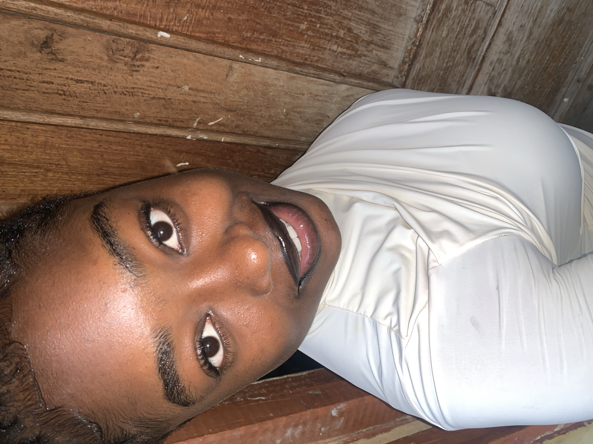 The media file shows a close-up, angled shot of a person lying down with their head tilted upwards. The subject has dark skin, dark eyes, and dark, curly hair. They are wearing a white, long-sleeved shirt. Their mouth is open, showing teeth, and their eyes are wide open. The background consists of horizontal wooden planks, with some visible texture and grain. There are faint white specks on the wood, possibly paint. The lighting appears to be artificial and casts some shadows, creating a somewhat dim atmosphere. The perspective is from above and to the side of the subject. No text is visible in the image. The scene appears to be indoors, possibly in a rustic or unfinished space due to the visible wooden structures. Given the location context of Rafin Par, Nigeria, this could be a domestic setting or a common area within a building. The emotion conveyed by the subject's facial expression is one of surprise or excitement.