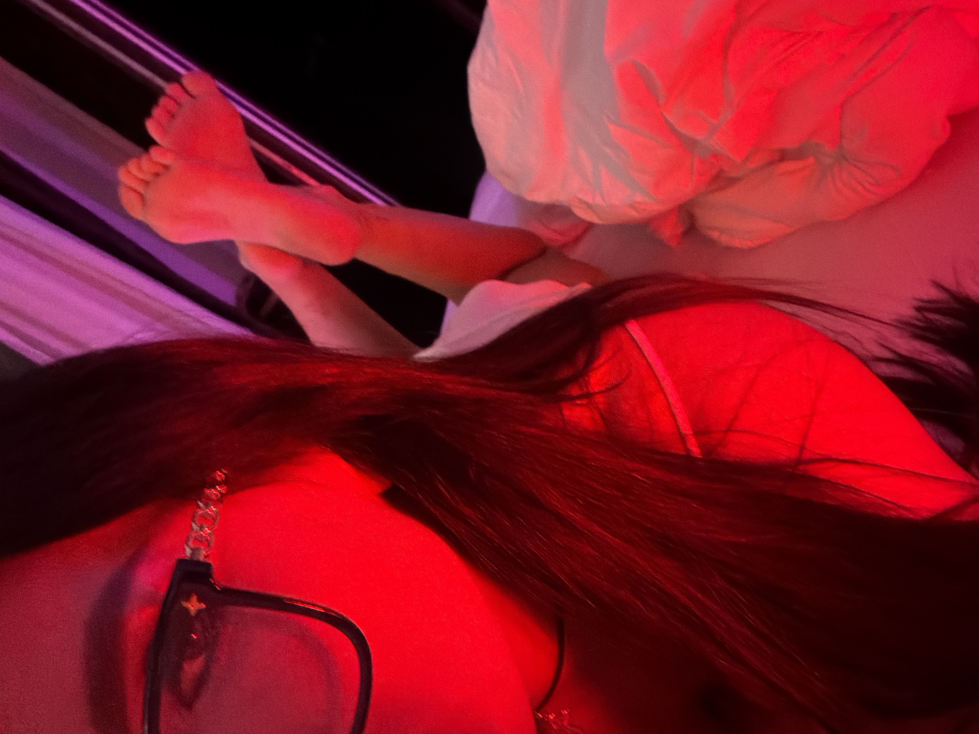 A person is depicted relaxing indoors in Boreti, Montenegro, bathed in dramatic red and purple ambient lighting. The shot is taken from a high angle, looking down. In the foreground, the person's upper body is visible, characterized by long, dark hair that flows around their head and shoulders. A pair of black-framed glasses, adorned with a silver chain, rests on the left side of their head. A distinct four-petal flower emblem, reminiscent of the Louis Vuitton monogram, is visible on the temple of the glasses. In the background, the person's bare feet are crossed and slightly elevated, resting on a striped surface that appears to be a hammock or striped bedding. A white blanket or garment covers part of their lower body, reflecting the intense red light. The overall atmosphere is intimate and tranquil, suggesting an evening or nighttime setting.