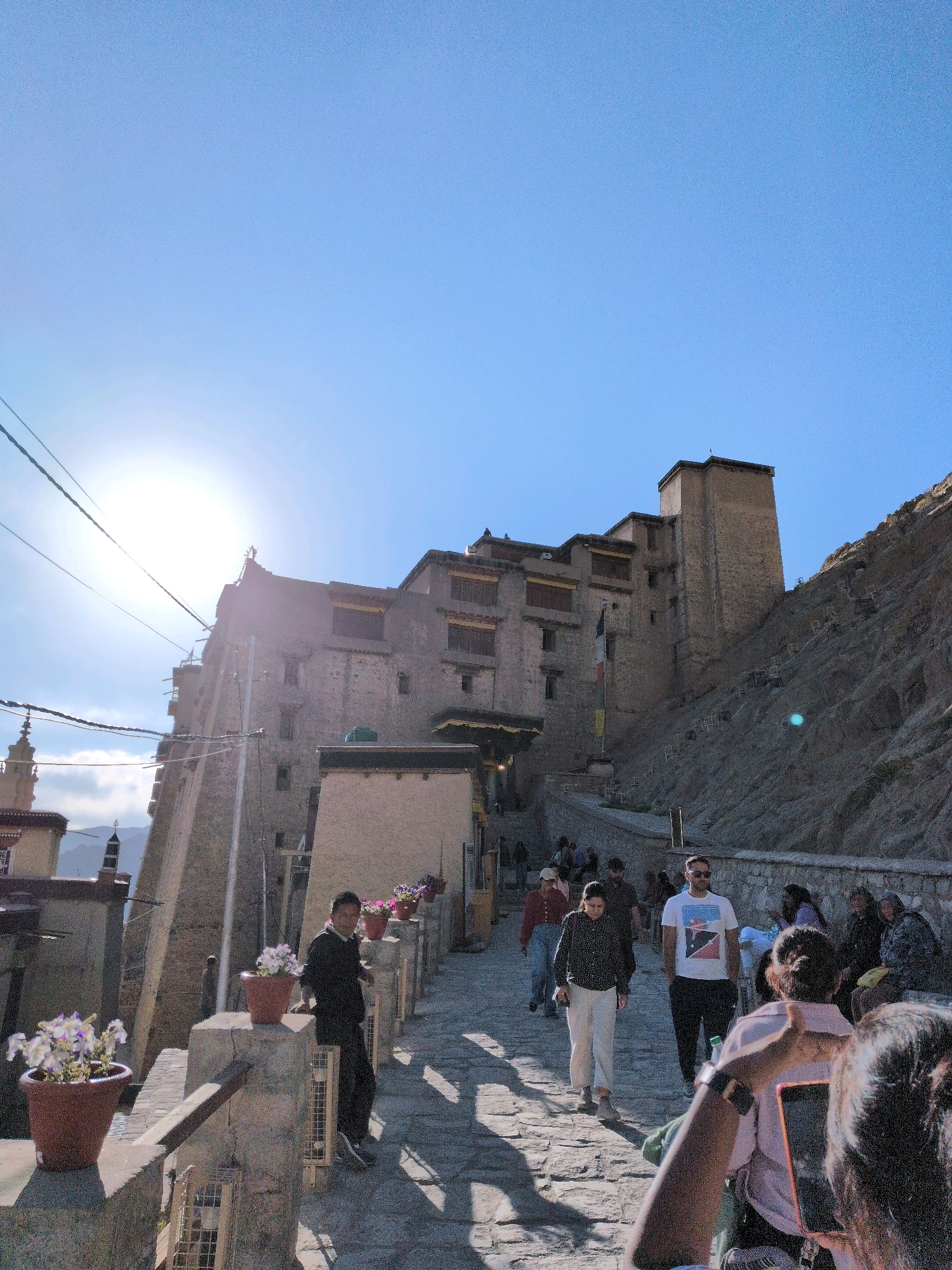 The image captures a view of the Leh Palace in Leh, India, on a sunny day. The palace, a towering structure built into a mountainside, dominates the background. Its multi-story, weathered stone facade is partially obscured by the bright sun, which creates lens flare and casts long shadows.
In the foreground, a stone pathway slopes upwards towards the palace. Several people are walking along this path and gathered in small groups. Some are dressed casually in t-shirts, pants, and jackets, while others appear to be dressed in more traditional attire. A person in the lower right corner is holding up a smartphone, seemingly taking a picture. Potted plants with white and pink flowers are placed along the pathway, adding a touch of color. Electrical wires crisscross the sky, and a minaret-like structure is visible in the distance on the left. The overall scene depicts a popular tourist destination, with visitors exploring the historical site under a clear blue sky.