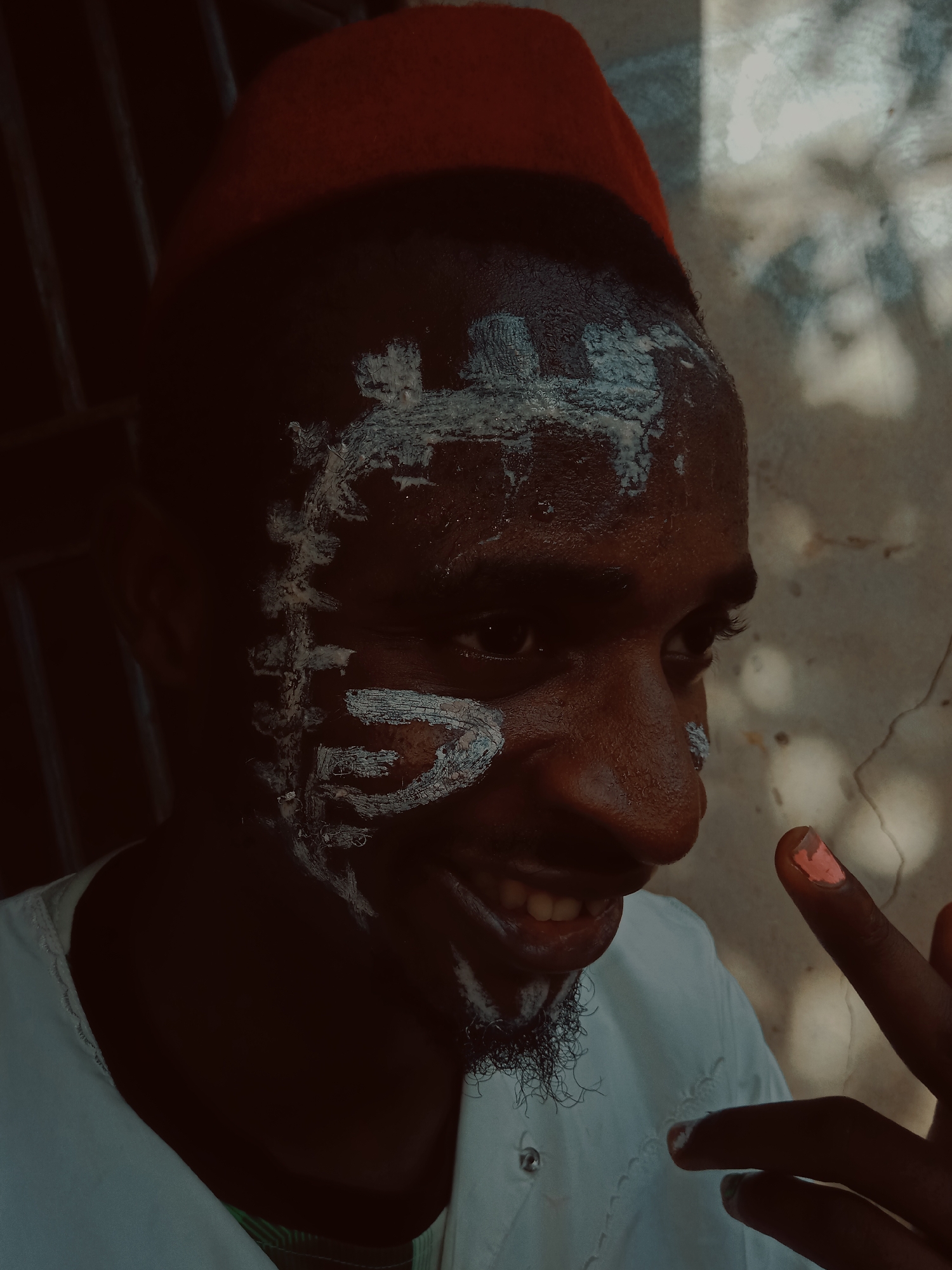 The image depicts a person with dark skin wearing a red hat and white face paint in a semi-obscured setting. The individual is smiling with teeth visible and gesturing with their right hand, where the index fingernail appears to be painted a light color. A white garment is visible on the person's upper body. The background is indistinct but seems to be a wall with some sort of light pattern. The location is specified as the city of Unknown, Unknown.