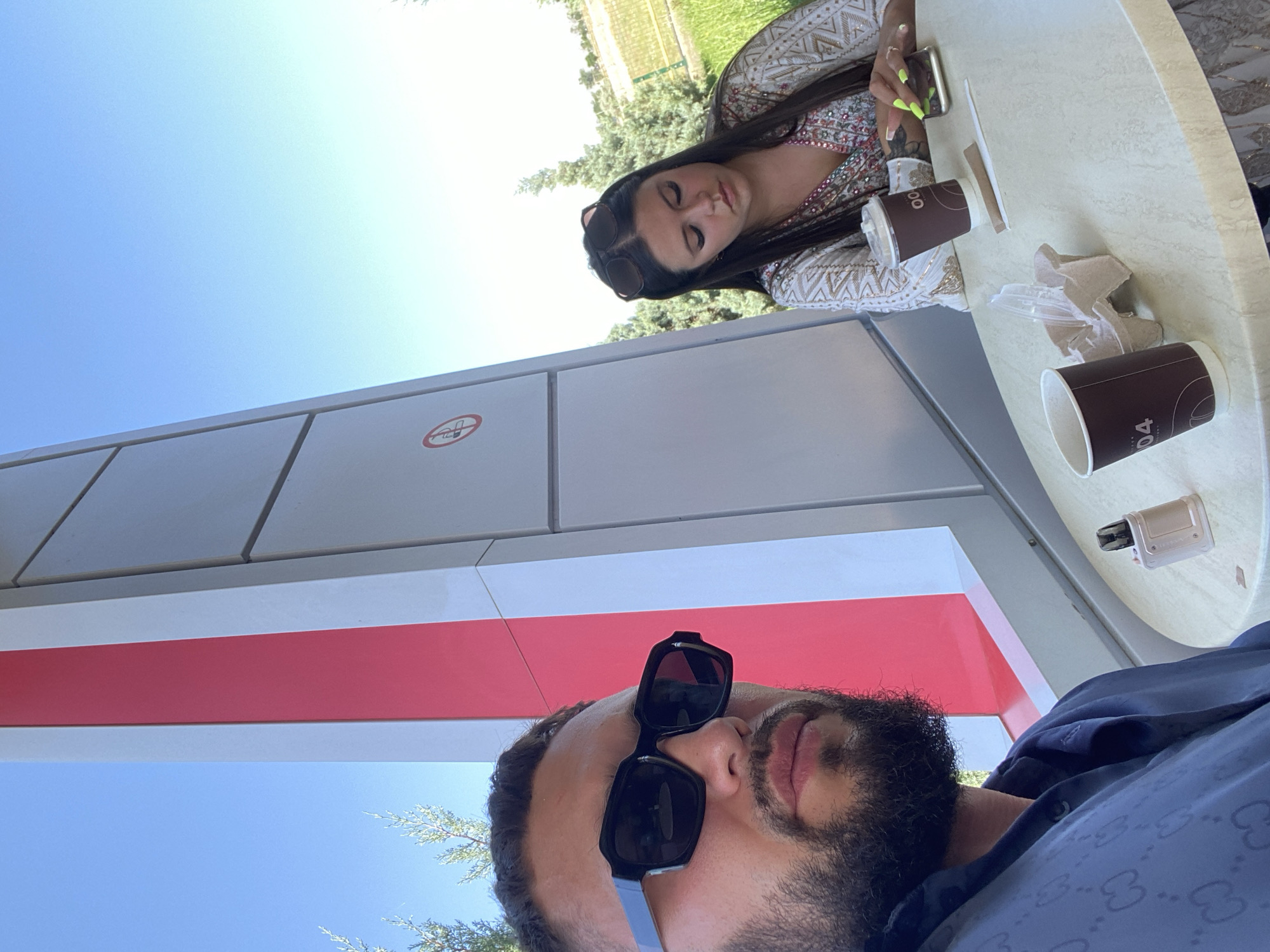 Two individuals are seated at a circular table outdoors. In the foreground, a man wearing black sunglasses and a dark blue patterned shirt is in profile, looking to his left. To his right, a woman with dark hair, wearing a white and gold embroidered top, looks down at her phone. Two disposable coffee cups and a vaping device are on the table. Behind them, a large, modern structure with red and white panels stands against a clear blue sky. The surrounding area appears to be a park or green space with trees and grass. The scene is likely located in the city of Korkmaskala, Russia, as suggested by the image context.