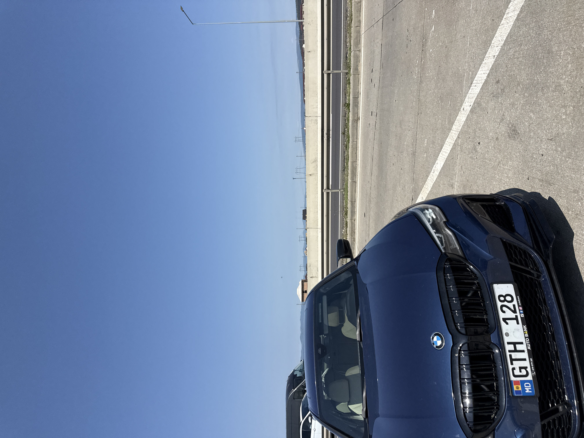 The image captures a blue BMW parked on a roadside in Sanpetru, Romania, under a clear blue sky. The car, with its license plate reading "GTH 128," is positioned in the foreground, facing the viewer. Behind the car, a concrete barrier separates the roadway from a distant landscape featuring power lines and buildings, suggesting an urban or semi-urban environment. The road surface is asphalt with visible white lane markings. To the left of the BMW, other vehicles, including a dark-colored van and a white car, are partially visible, indicating a traffic flow or staging area. The overall scene is bright and appears to be taken during daylight.