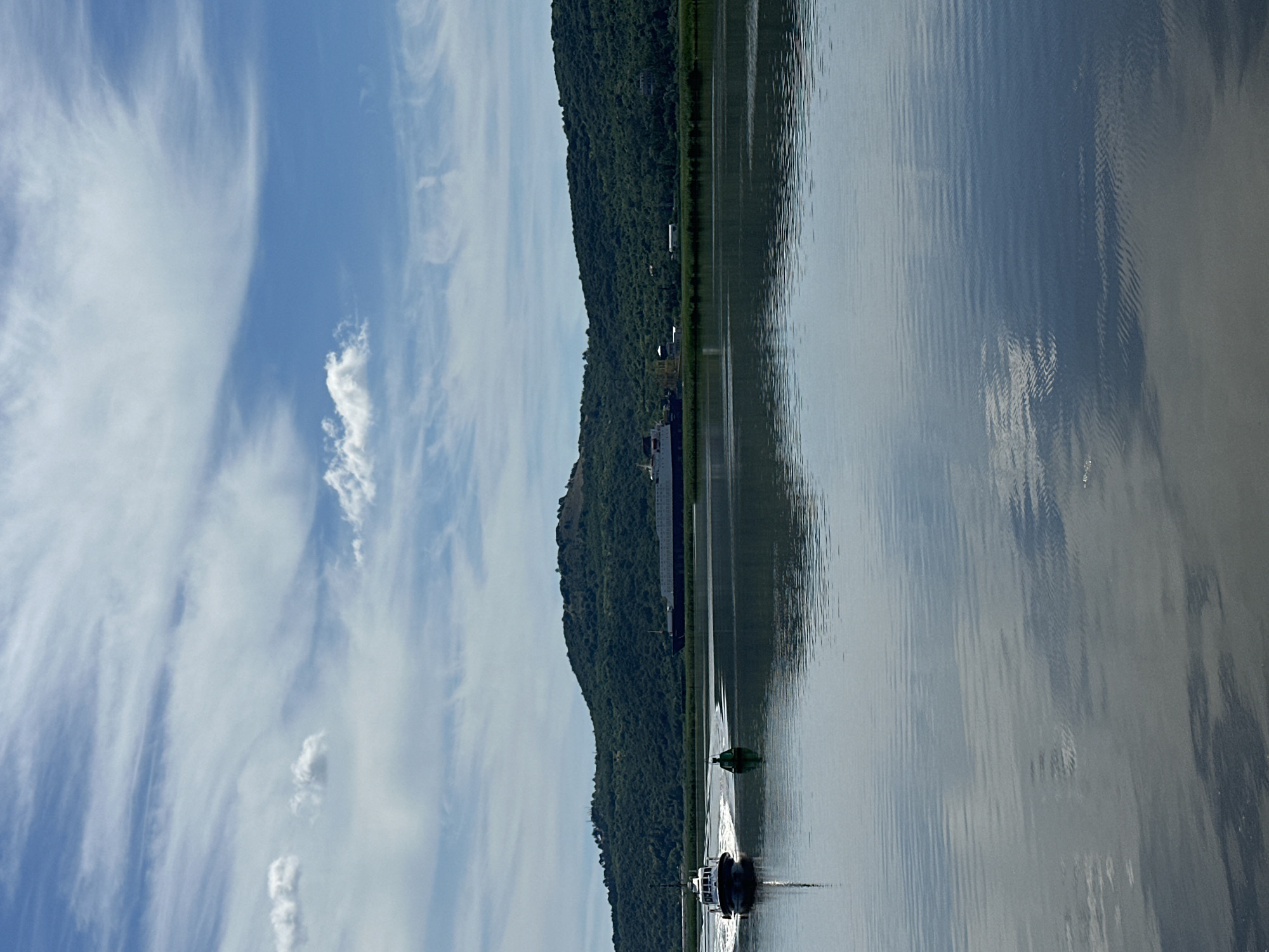 A low-angle, wide shot captures a serene scene on a body of water with a verdant, tree-covered hill in the background under a partially cloudy blue sky. In the foreground, the water's surface is calm, with ripples and reflections of the sky and land. On the left side of the frame, a white boat with a dark trim and a single figure at the helm is moving away from the viewer, leaving a white wake. Adjacent to the boat, a small, green, cylindrical buoy floats. Further in the distance, a long, dark ship is docked or anchored near the shore, parallel to the waterline. The hill behind the ship is densely forested, with a few buildings visible near its base and a faint structure atop the hill. The overall impression is one of peaceful coexistence between human activity and nature.