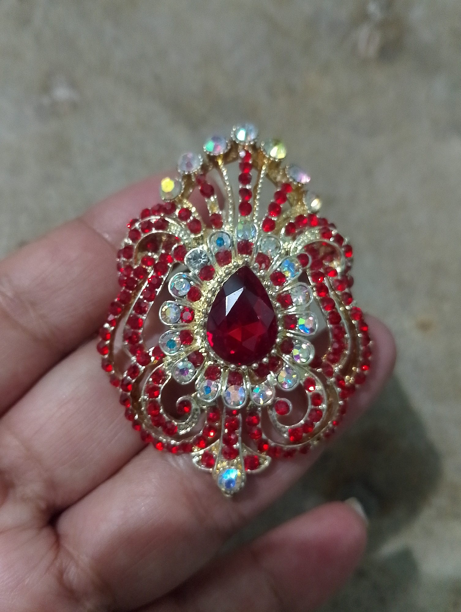 In Serbelawan, Indonesia, a hand holds a circular brooch adorned with red and clear rhinestones and a large, teardrop-shaped red gem at its center. The brooch features intricate gold-colored metalwork resembling swirling vines and floral motifs. The background is a textured, neutral-colored surface, possibly a wall or tabletop. The lighting suggests an indoor setting.