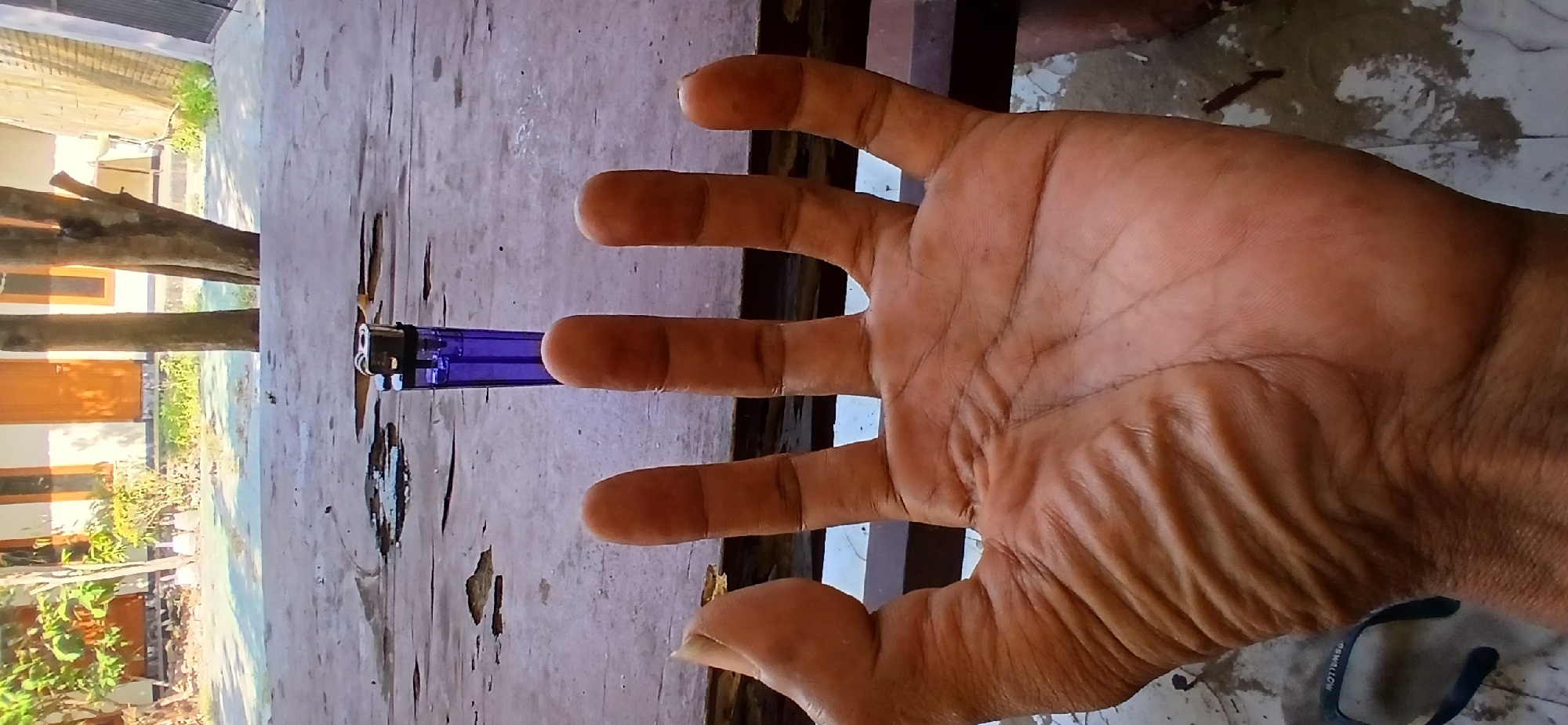 A close-up shot shows a person's left hand and palm resting on a weathered wooden surface. The palm is wrinkled and tanned, with visible lines. Four fingers are extended upwards, with the thumb bent towards the palm. Behind the hand, on the wooden surface, rests a purple plastic lighter. The background shows a blurred outdoor scene with trees, buildings with orange doors, and a sandy ground. The image appears to be taken in Pejaten, Indonesia, indicated by the specific details of the environment.