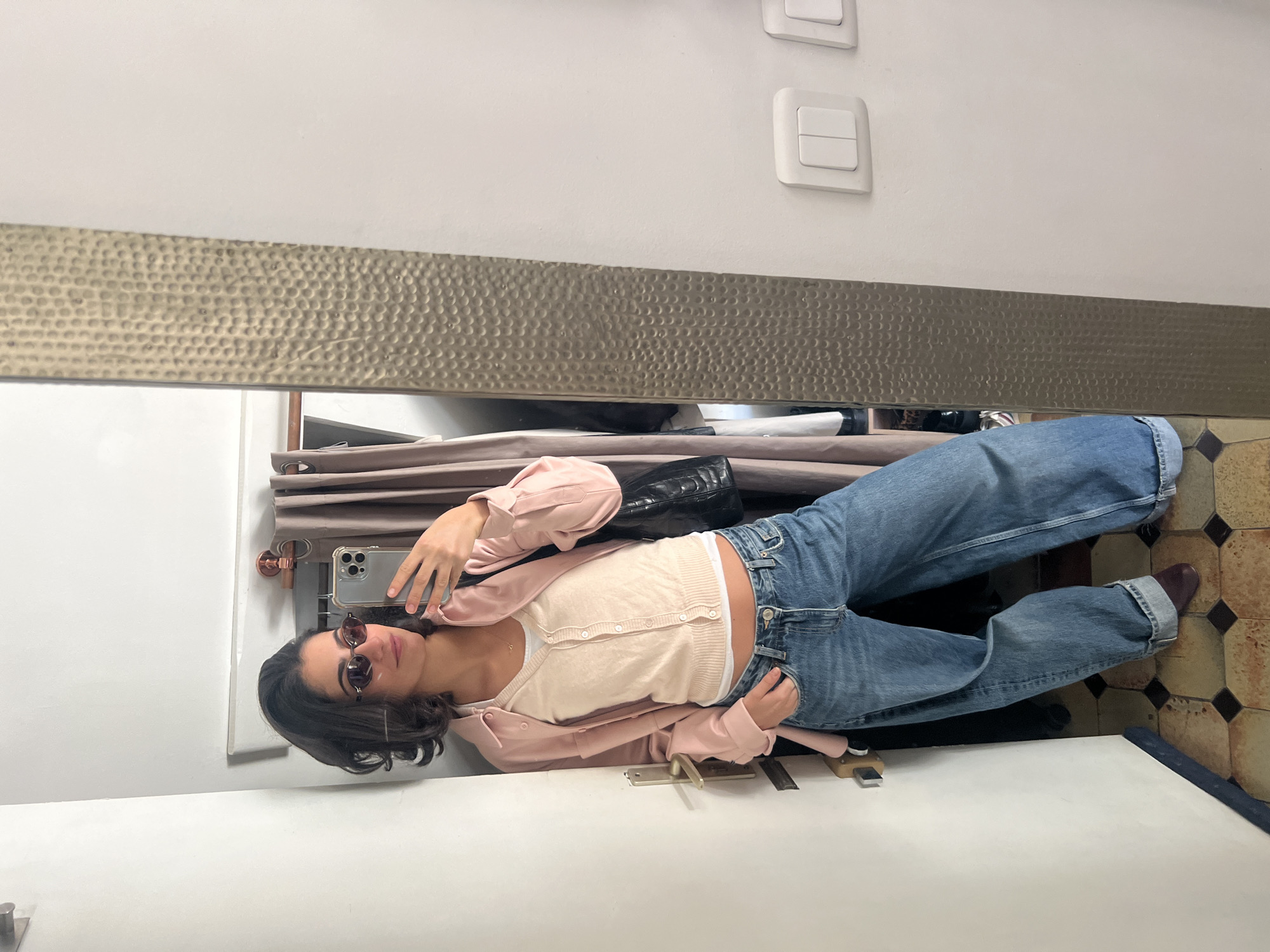 The image is a mirror selfie of a young woman in Paris. She wears sunglasses, a pink cardigan over a light-colored, button-up shirt, and blue jeans. Her hair is dark and curly. She is holding a smartphone to take the picture. Behind her in the reflection is a curtain and a black bag. The wall above the mirror has a textured band and two light switches. The floor is tiled in a geometric pattern. The woman appears to be standing in a room, possibly a bathroom or dressing area, within a Parisian building.