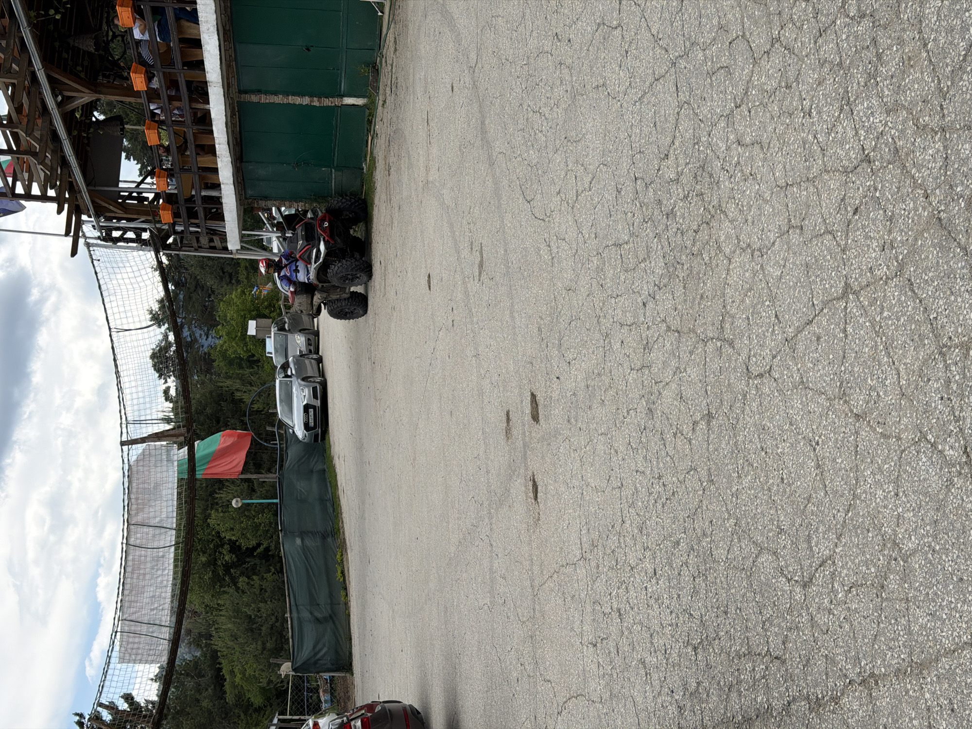 The image captures an outdoor scene, likely a recreational area or adventure park, set in a natural environment consistent with locations like Цигов чарк, Bulgaria, as indicated by the prominent Bulgarian flag and vehicle license plate. The foreground features a cracked asphalt or paved surface, exhibiting widespread alligator cracking, with several dark stains or patches.
On the right side of the image, a two-story wooden structure is visible. Its lower level has a dark green facade with two closed green doors, while the upper level is an open wooden balcony or terrace. The balcony railing supports several orange rectangular containers. Indistinct figures are seated at tables on this upper level, engaged in leisure activities. At the very top of the building, multiple flags are flying, including what appears to be another Bulgarian flag.
To the left of the building and further back, a person wearing a red and white helmet and a blue and white long-sleeved top is seated on a black and red All-Terrain Vehicle (ATV) with large knobby tires. The ATV is positioned on the paved surface, facing slightly to the left. Behind the ATV, a silver Audi A4 sedan (Bulgarian license plate PB 6695 PE) is parked. Another silver car is partially visible behind the Audi.
In the mid-ground, a dark green privacy fence or screen runs horizontally. Behind this fence, a large Bulgarian flag (white, green, red horizontal stripes) is flying from a pole. Further back, spanning across the upper mid-ground, is a suspended rope bridge or net course, constructed from mesh netting and wooden elements, suggesting an adventure activity. The entire background is filled with dense green trees under a partly cloudy sky. A white, possibly canine-shaped, object is faintly visible near the left end of the green fence.