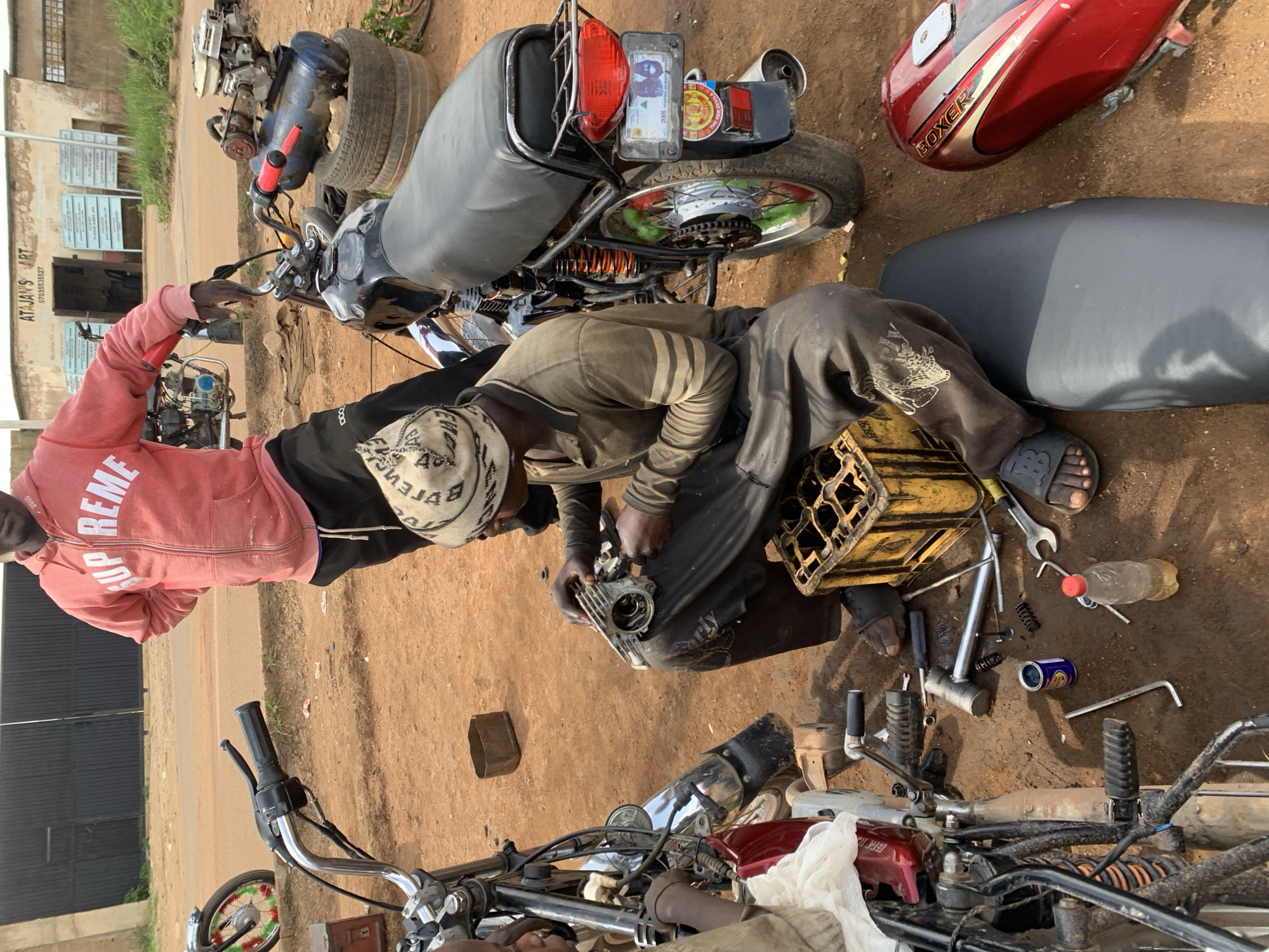 In Barakin Lerawi, Nigeria, two men are working on motorcycles outside a garage. The man on the right, wearing a pink hooded sweatshirt and black pants, is standing. The man on the left, wearing a brown long-sleeved shirt and black pants, is seated, holding a motorcycle engine part. Various tools, including wrenches, screwdrivers, and an air compressor, are scattered around them on the dusty ground. Two motorcycles, one red and one black, are present, one of which is partially disassembled. A yellow plastic crate is positioned near the seated man. The background shows a building with signage and a closed black gate.