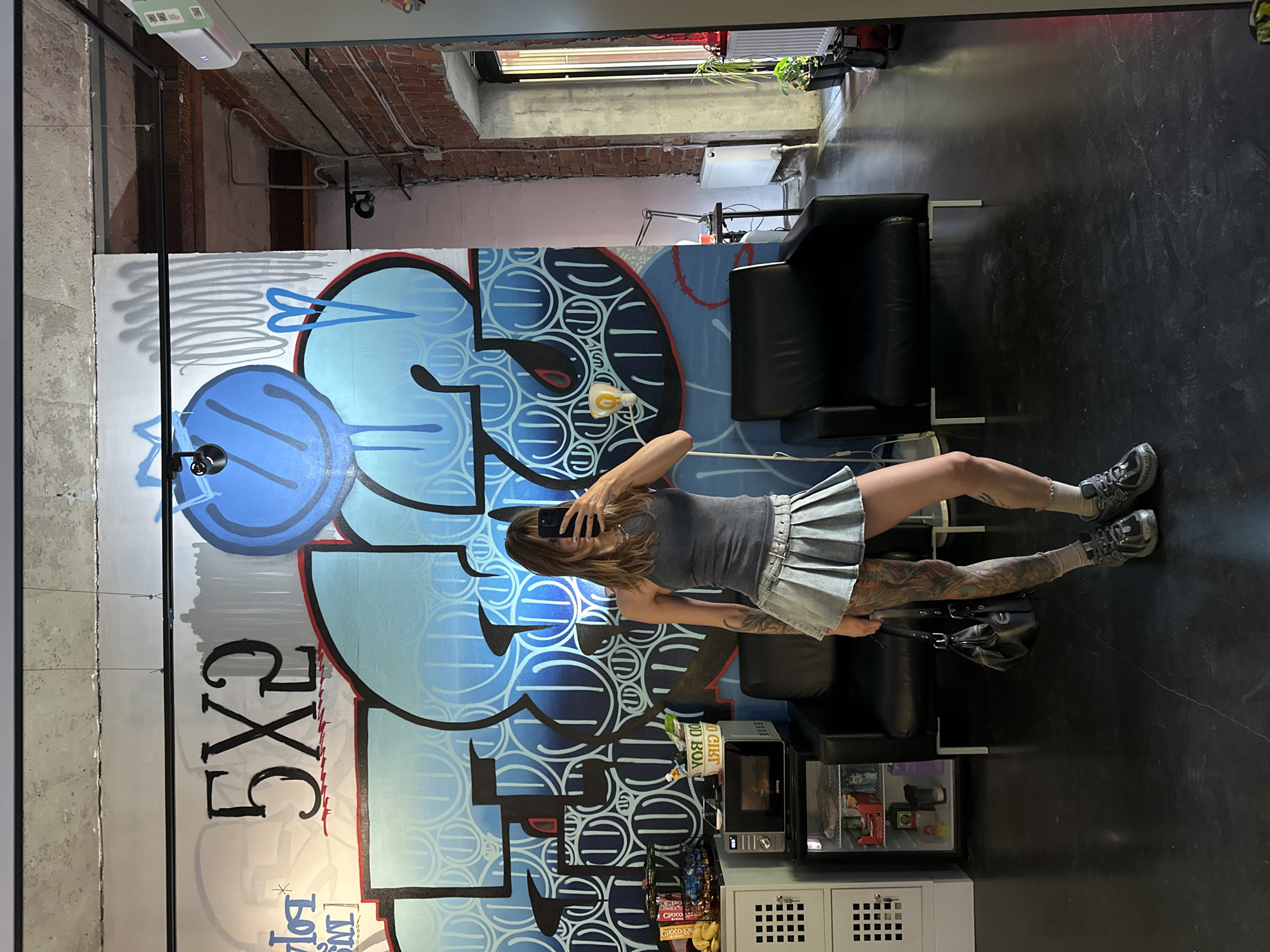A woman stands in a room featuring a wall covered in graffiti. The graffiti includes blue and black designs, numbers, and lettering. The woman is holding a phone, presumably taking a picture of herself in front of the mural. She wears a grey tank top and a pleated denim skirt. Her legs are tattooed. Two black leather chairs are in the room. A small microwave and mini-fridge are next to the chairs. The floor is black and marbled. The location is Moscow, Russia.