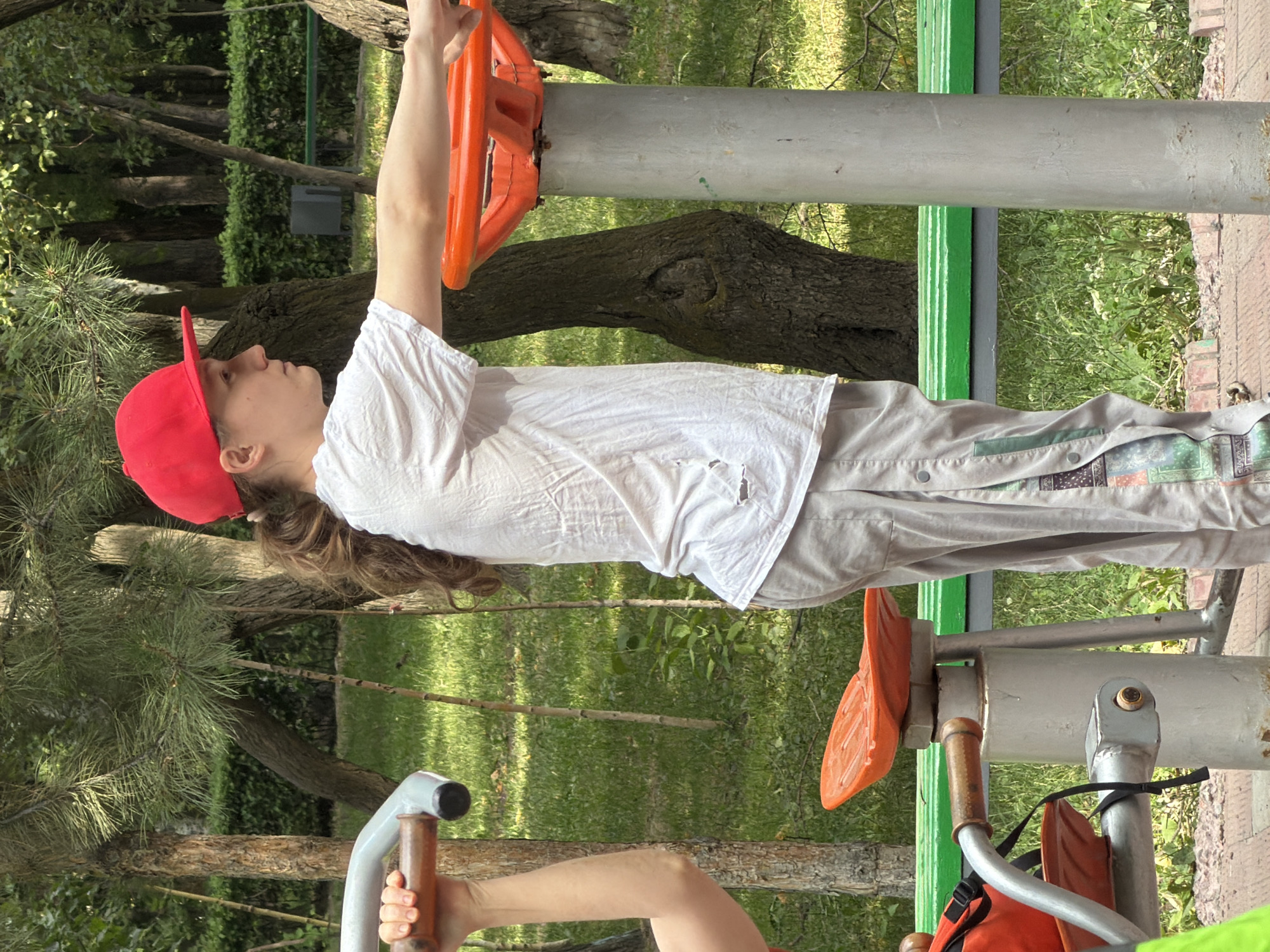 A young person wearing a red baseball cap and a white t-shirt is standing outdoors at a public park exercise station. They are facing right in profile and appear to be using an exercise machine with an orange steering wheel. The person's left arm is extended, and their right arm is bent at the elbow, with their hand on the orange wheel. They are wearing light-colored pants. To the left, a portion of another person's arm and hand are visible, holding onto a metal bar of a different piece of exercise equipment. The background consists of trees, green grass, and a green park bench. The ground is paved with brick-like material.