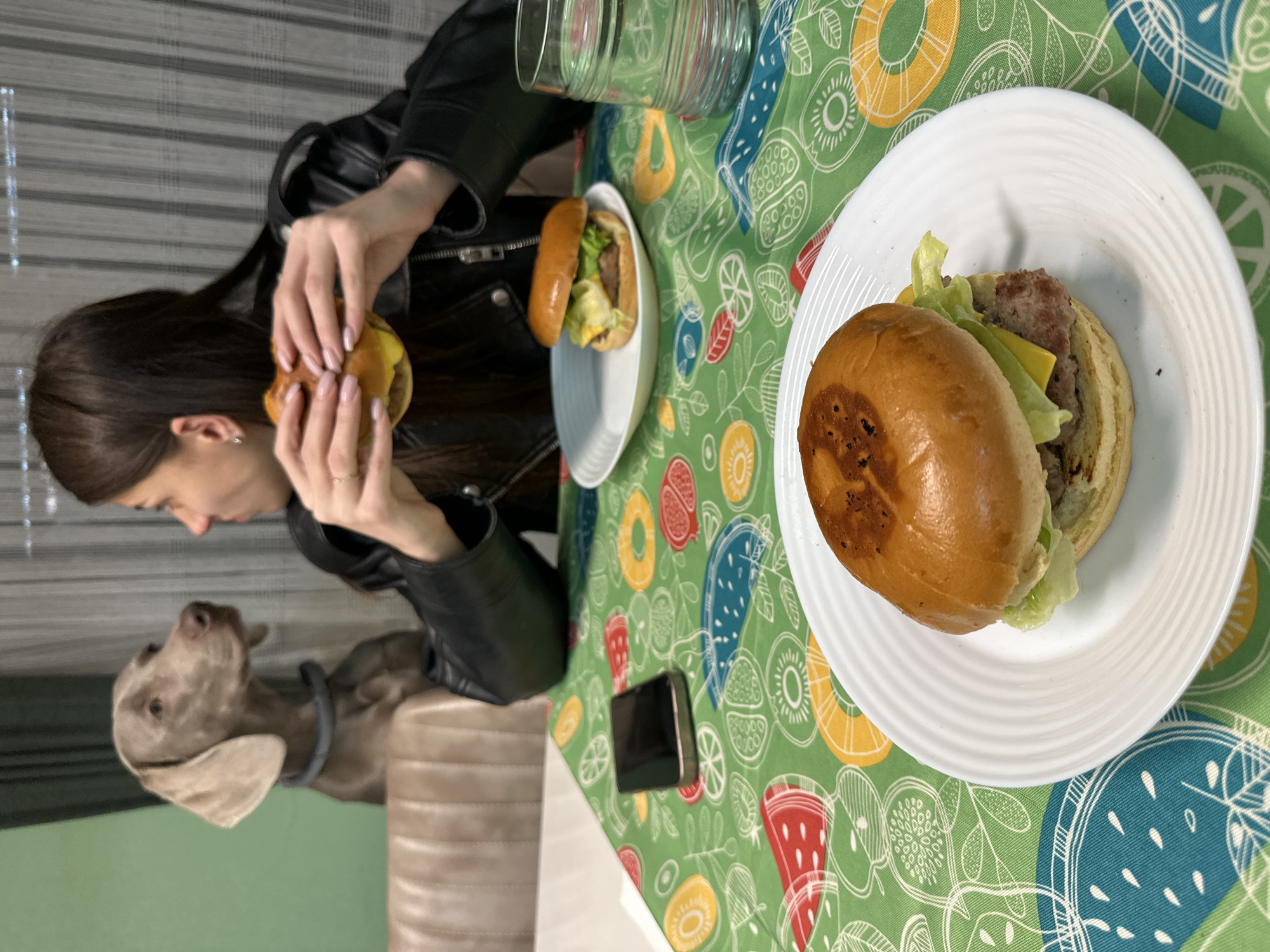 The image depicts a woman in a black leather jacket holding a hamburger with both hands, seemingly about to eat it. A Weimaraner dog is seated next to her, looking up towards the woman. On the table is a green tablecloth with a colorful fruit pattern. There are two hamburgers on white plates, a glass pitcher, and a black smartphone on the table. The scene appears to be indoors. The precise location in Plovdiv, Bulgaria, cannot be determined from this image alone.