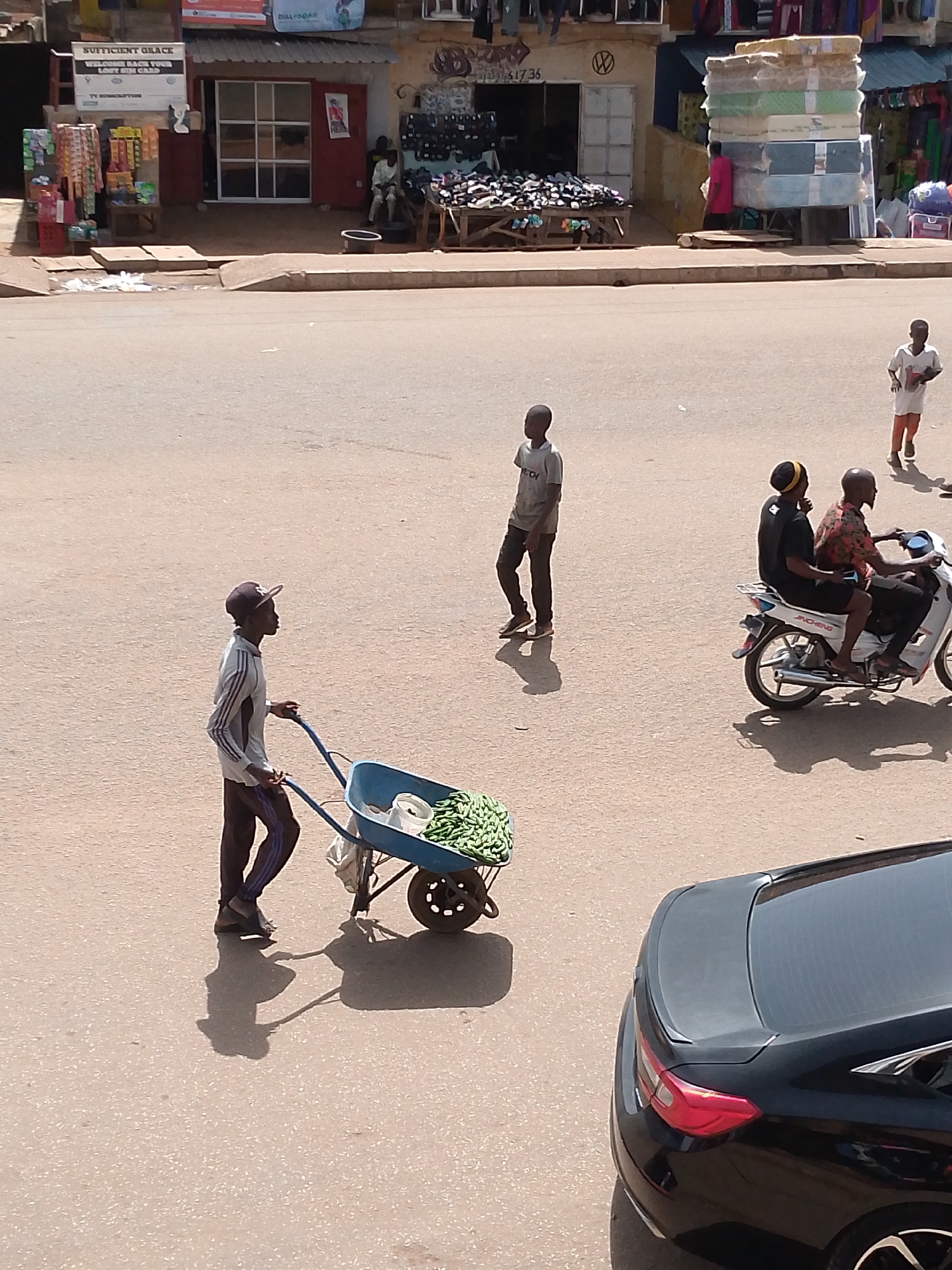 A young boy pushes a wheelbarrow filled with green produce down a paved street. He wears a striped long-sleeved shirt and dark pants. In the background, two men ride a motorcycle, and another young boy stands by himself. A black car is parked in the foreground. Buildings with shops are visible in the distance.