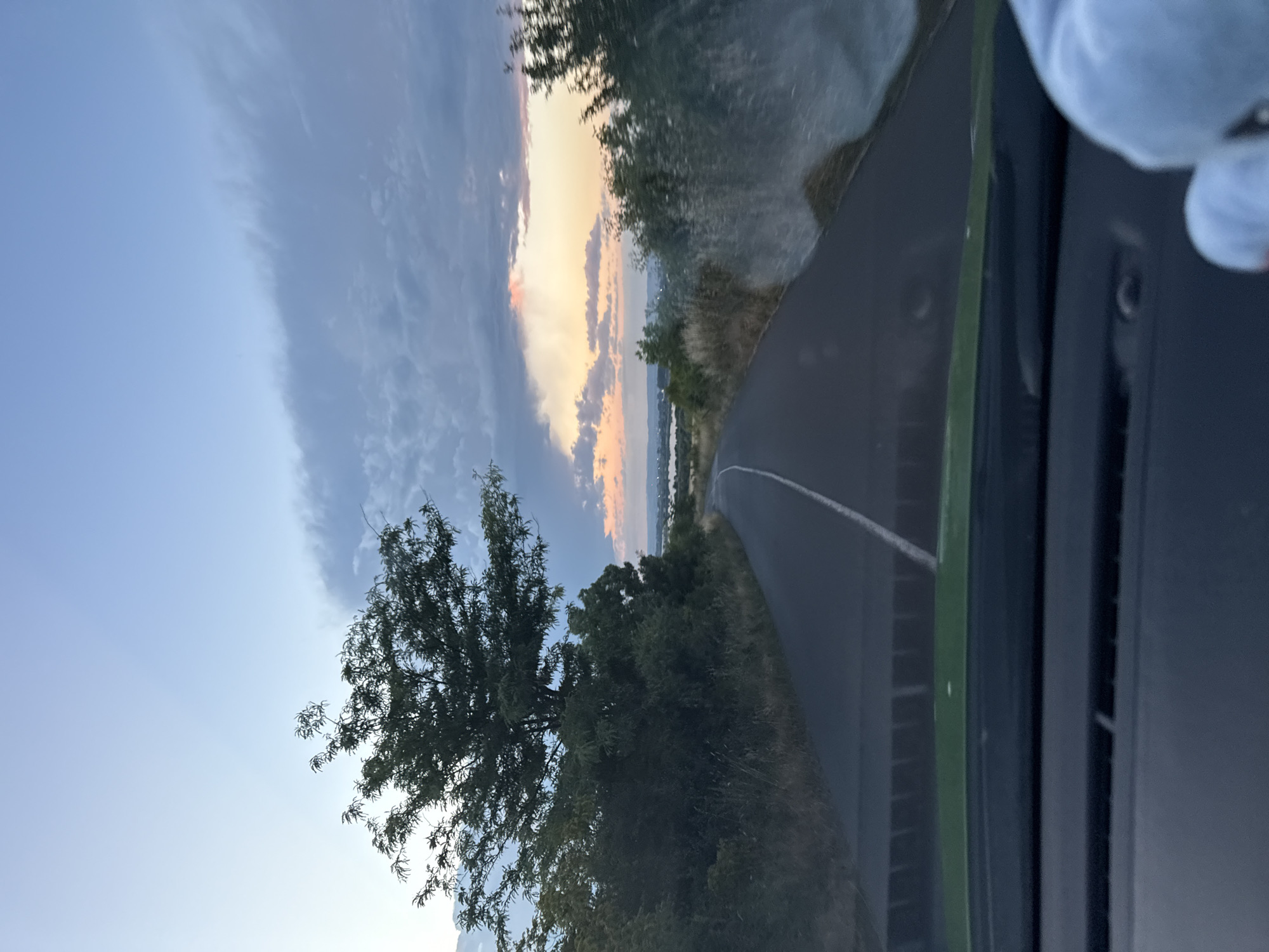 A lone asphalt road with a white line down the middle curves through a landscape under a twilight sky. The sky transitions from a pale blue at the top to hues of orange and pink near the horizon, where scattered clouds are visible. On the left side of the road, a large, leafy tree stands amidst dense green foliage and dry grass. On the right, more greenery and trees are present, some appearing blurred, possibly due to motion. In the distance, a body of water and hints of buildings can be seen. The foreground shows the interior of a vehicle, with a dark dashboard and a portion of a green surface.