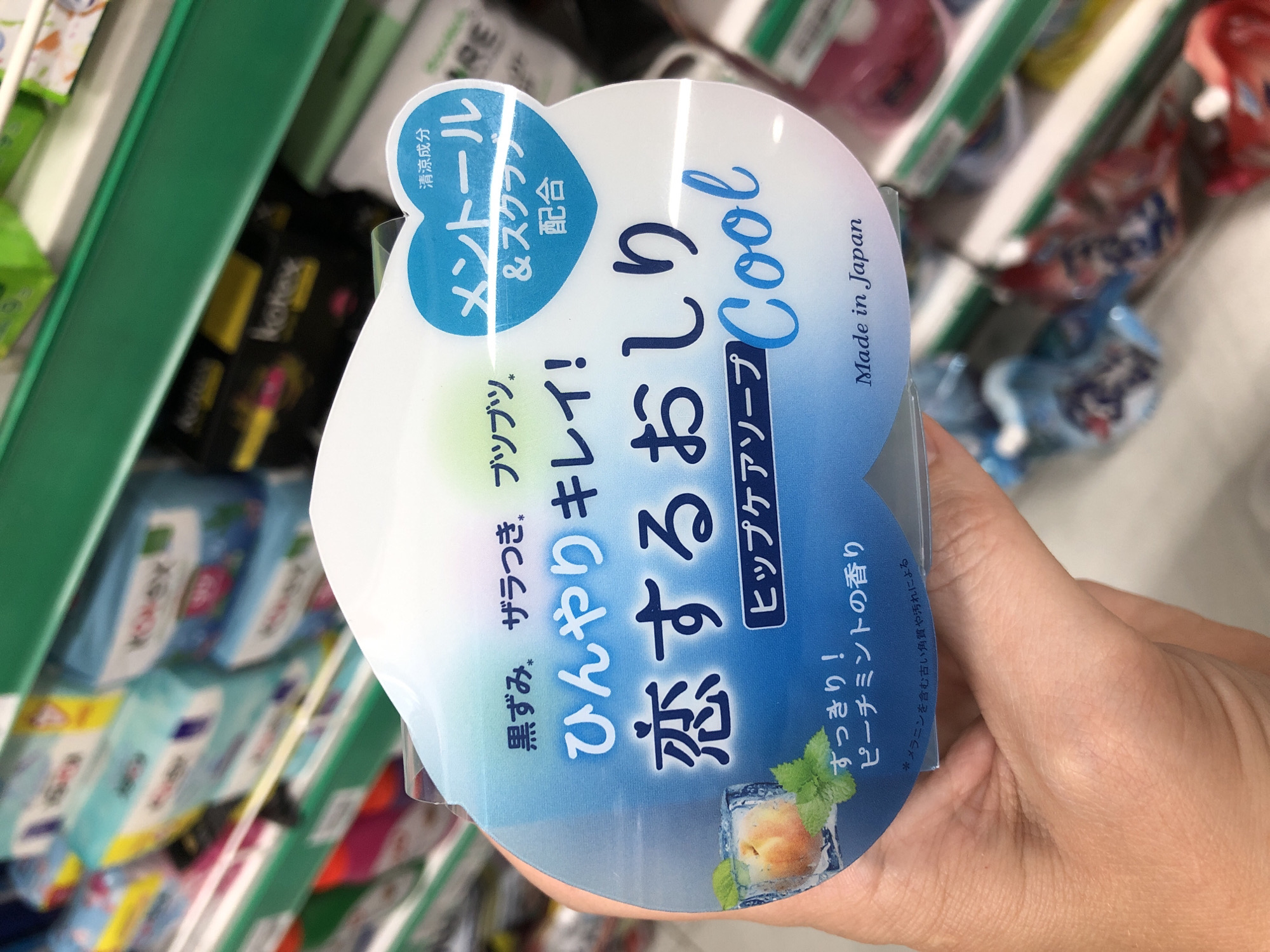The image shows a close-up of a hand holding a product with Japanese text. The product is likely a soap or skincare item, as indicated by the text "ヒップケアソープ" (Hip Care Soap) and the image of a peach with ice cubes. The packaging is predominantly white and blue, with a gradient effect. The text "メントール" (Menthol) and "Cool" suggest a cooling sensation. Other visible text includes "Made in Japan," "黒ずみ。ザラつき。ブツブツ。" (Blackheads, roughness, bumps.), "ひんやりキレイ!" (Cool and beautiful!), and "ピーチミントの香り" (Peach mint scent). The background appears to be a retail environment, with shelves filled with various products, suggesting the item is being displayed for sale. The lighting suggests an indoor setting, possibly a supermarket or drug store. The image is slightly tilted, and the focus is on the product and the hand holding it. The person's skin tone suggests it could be a person of Asian descent.