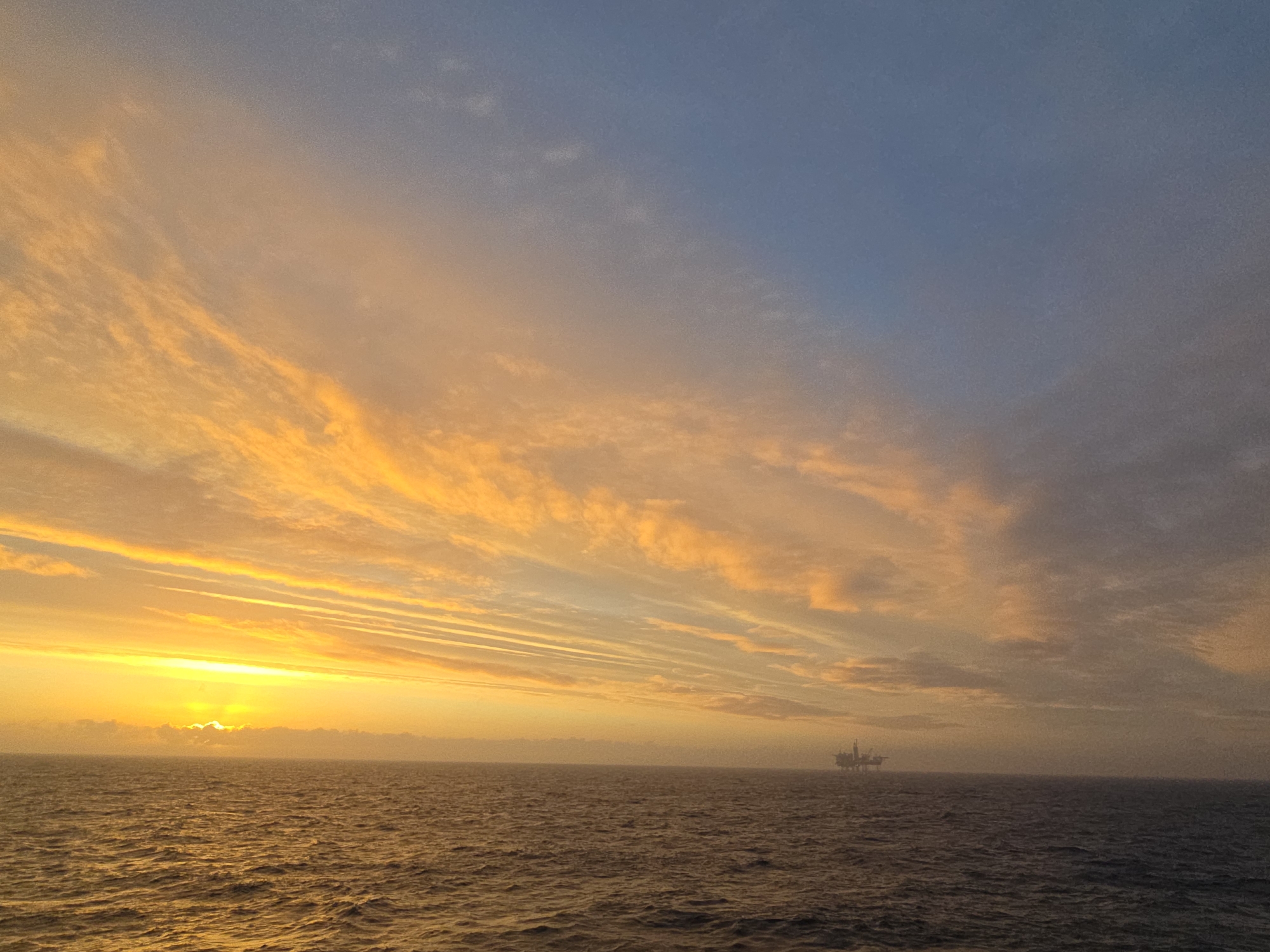A wide shot captures a body of water under a sky ablaze with color. The sun, obscured by clouds, casts a bright glow near the horizon, while the clouds above transition from orange to blue. An oil rig is visible in the distance on the horizon. The location is an unknown city.