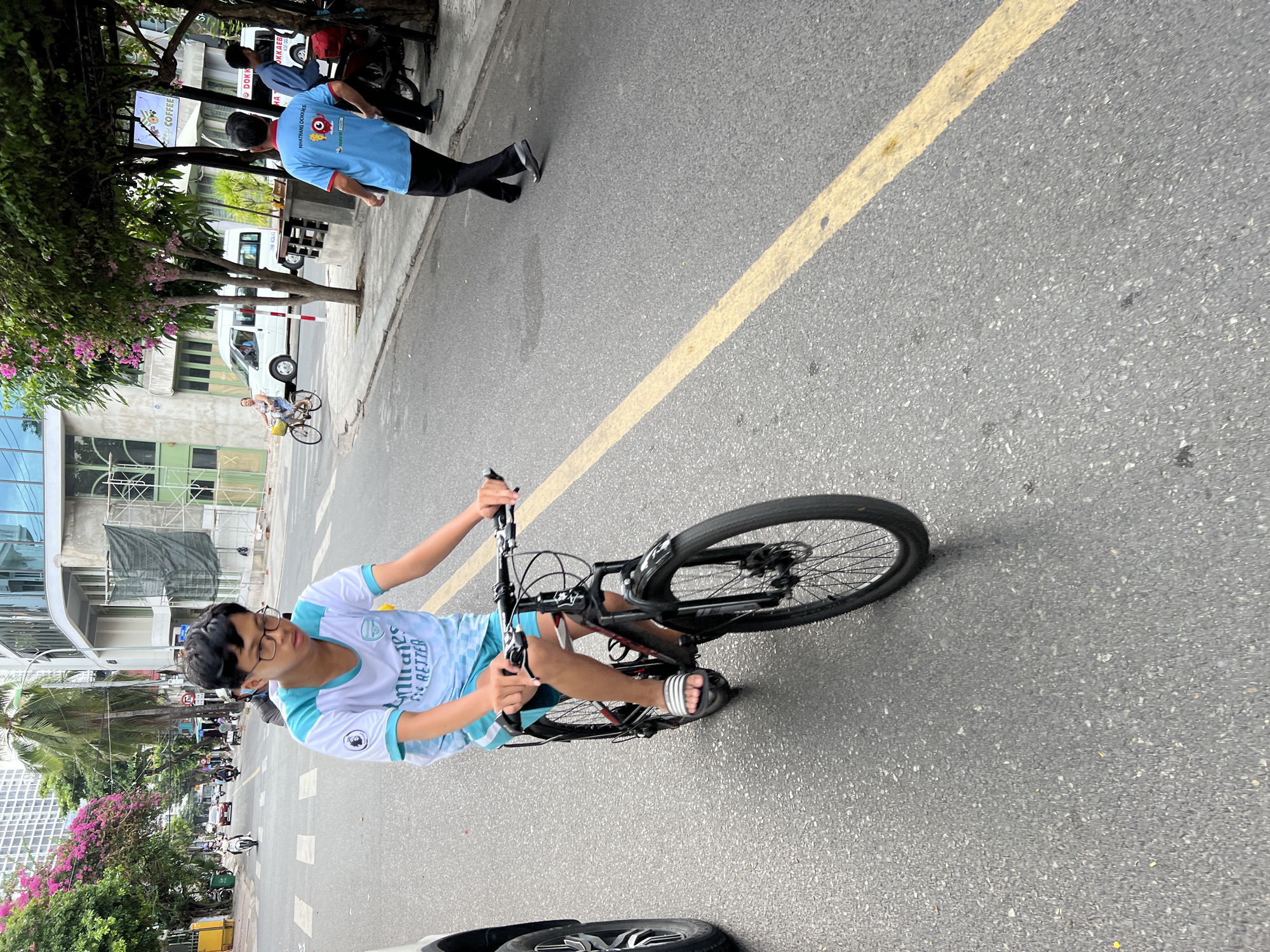A young male wearing glasses, a white t-shirt with blue trim, and blue shorts rides a black bicycle on a paved road in Nha Trang, Vietnam. The boy is wearing white and gray striped slides on his feet. The road has a yellow line marking the lane. In the background, there are trees with pink flowers, buildings, and other vehicles including motorcycles and cars. Two men in blue shirts and black pants are standing on the sidewalk to the right of the frame, next to a parked motorcycle. A white van is visible in the background.