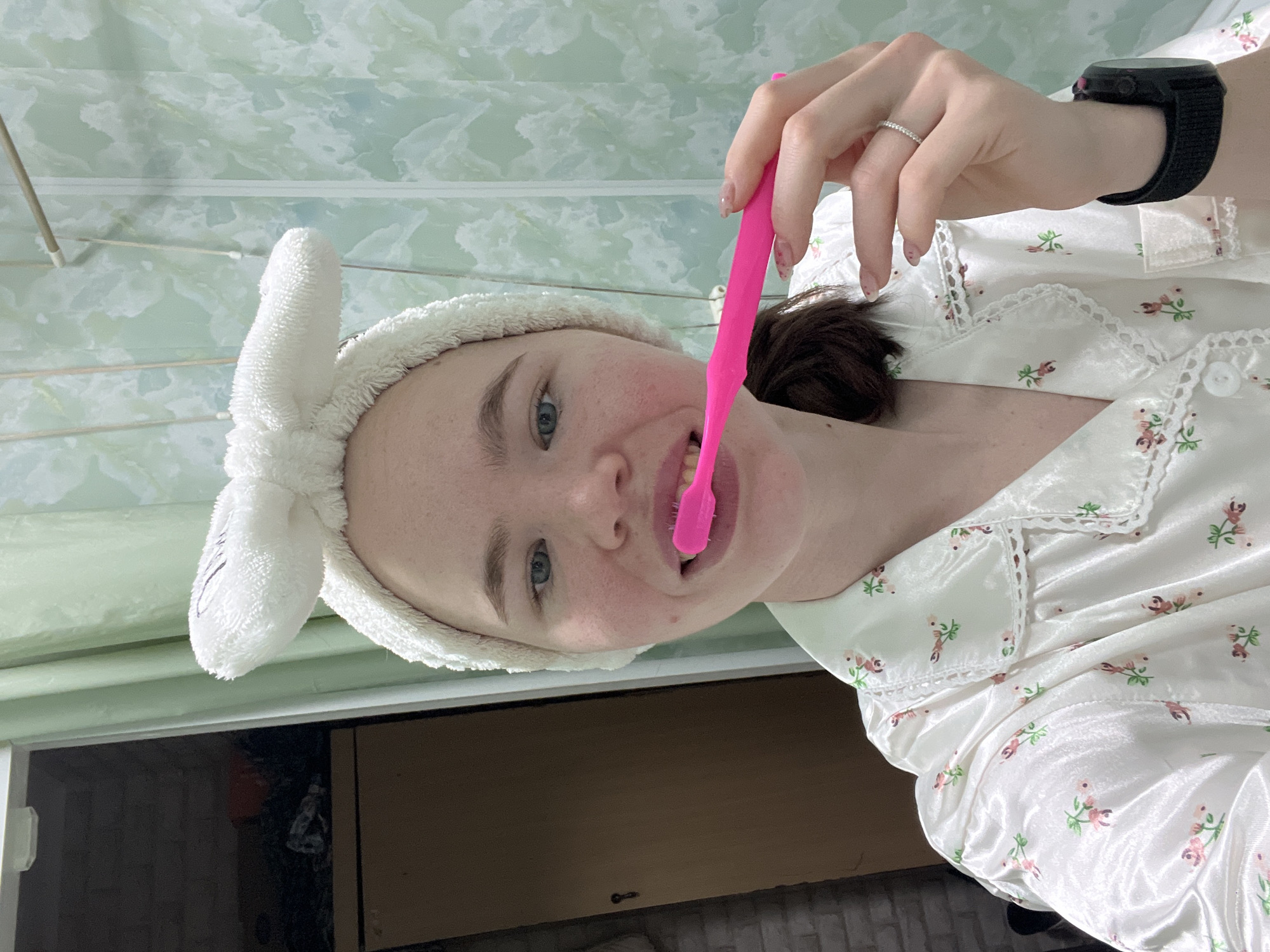 A woman is brushing her teeth. She is wearing a white bunny-ear headband and a white floral-patterned pajama top with lace trim. She has a pink toothbrush in her mouth and is holding it with her right hand, which has a ring on her finger. She is wearing a black smartwatch on her left wrist. The background includes a textured, light-green wall, a window, and a wooden cabinet. The location is Chaikovsky, Russia.