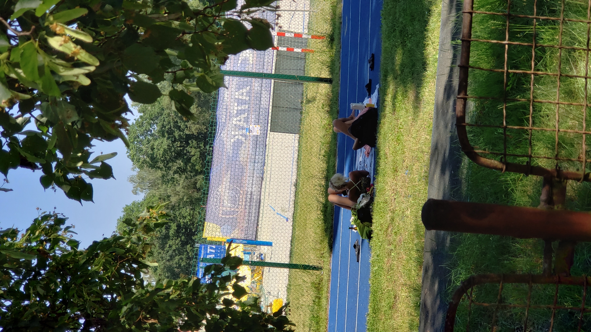 The image depicts a scene at a track and field facility, likely located in the city of Prešov, Slovakia. In the foreground, a rusty, dilapidated metal fence with square grid openings partially obscures the view. Beyond the fence, a blue athletic track runs horizontally across the frame. On the track, two individuals are lying down, seemingly relaxing or sunbathing. The person on the left has blonde hair and is wearing a patterned swimsuit, lying on their side. Next to them are several items, including a green bottle and a dark bag. The person on the right is wearing dark clothing and is lying on their back. They have a white bottle and a dark bag beside them. The track is bordered by a strip of green grass. In the background, there is a large banner with text and graphics, possibly advertising an event or sponsor. Behind the banner, there are buildings and a dense line of trees, suggesting an outdoor urban or suburban environment. The sky above is clear and blue, indicating a sunny day. The overall impression is one of a casual, recreational moment taking place at a sports venue.