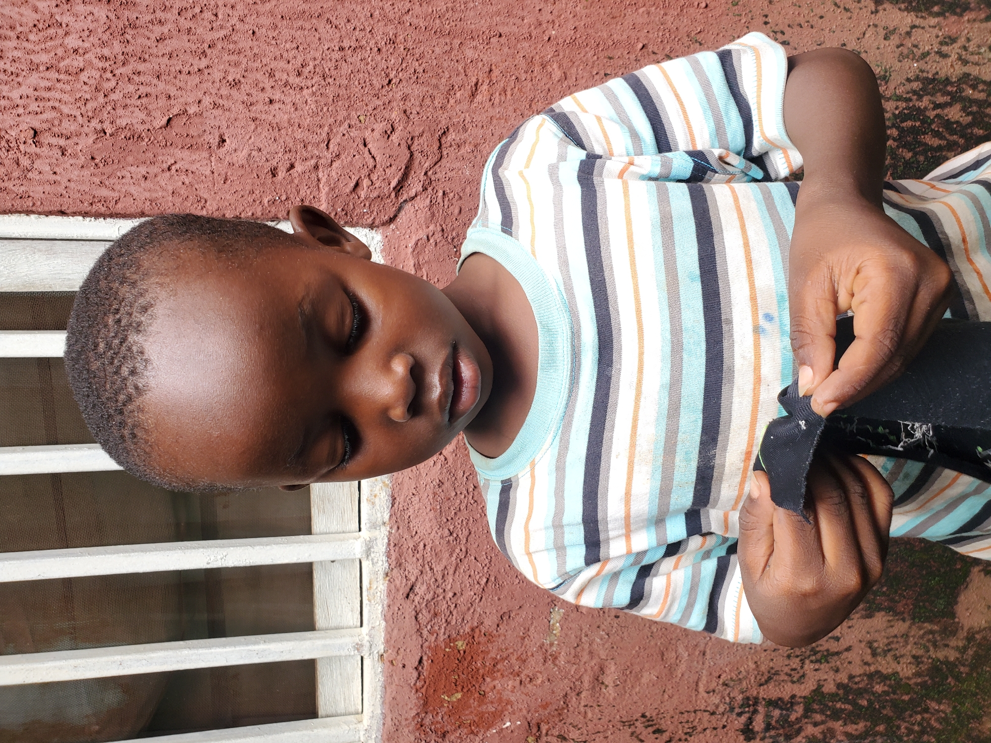 The image shows a young Black child, likely a boy, with his eyes closed and head tilted downwards. He is wearing a striped t-shirt with colors of blue, white, orange, and gray. His hands are engaged in inspecting a piece of black fabric, possibly a hem or a tear. The child appears to be outdoors, with a reddish-brown textured wall behind him and a white window grate to his left. The ground is visible below, with some vegetation. The lighting suggests it is daytime, possibly sunny, given the shadows and the brightness of the scene. The overall mood of the image is one of quiet contemplation or focus on the task at hand. There is no visible text in the image. The location context provided is Jos, Nigeria.